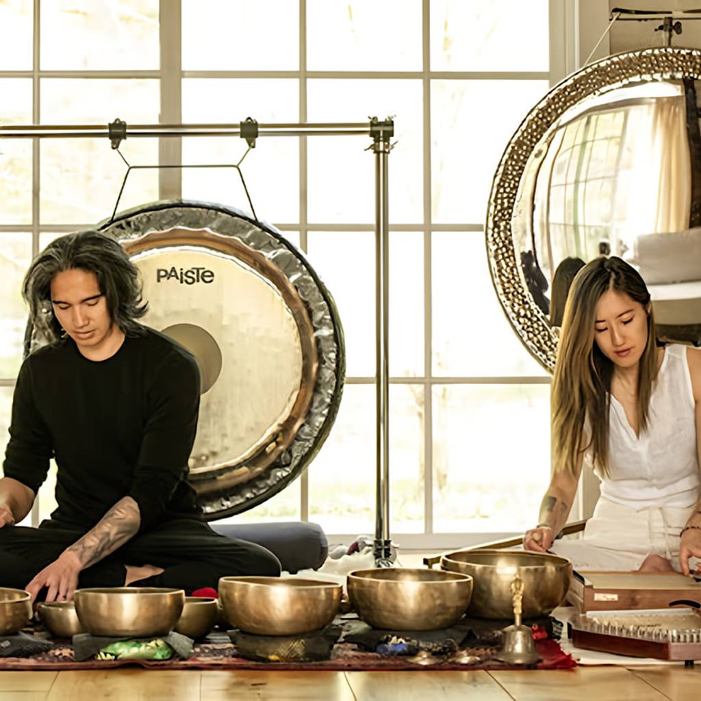 Two people sit among singing bowls, a large gong, and a stringed instrument in a bright room.
