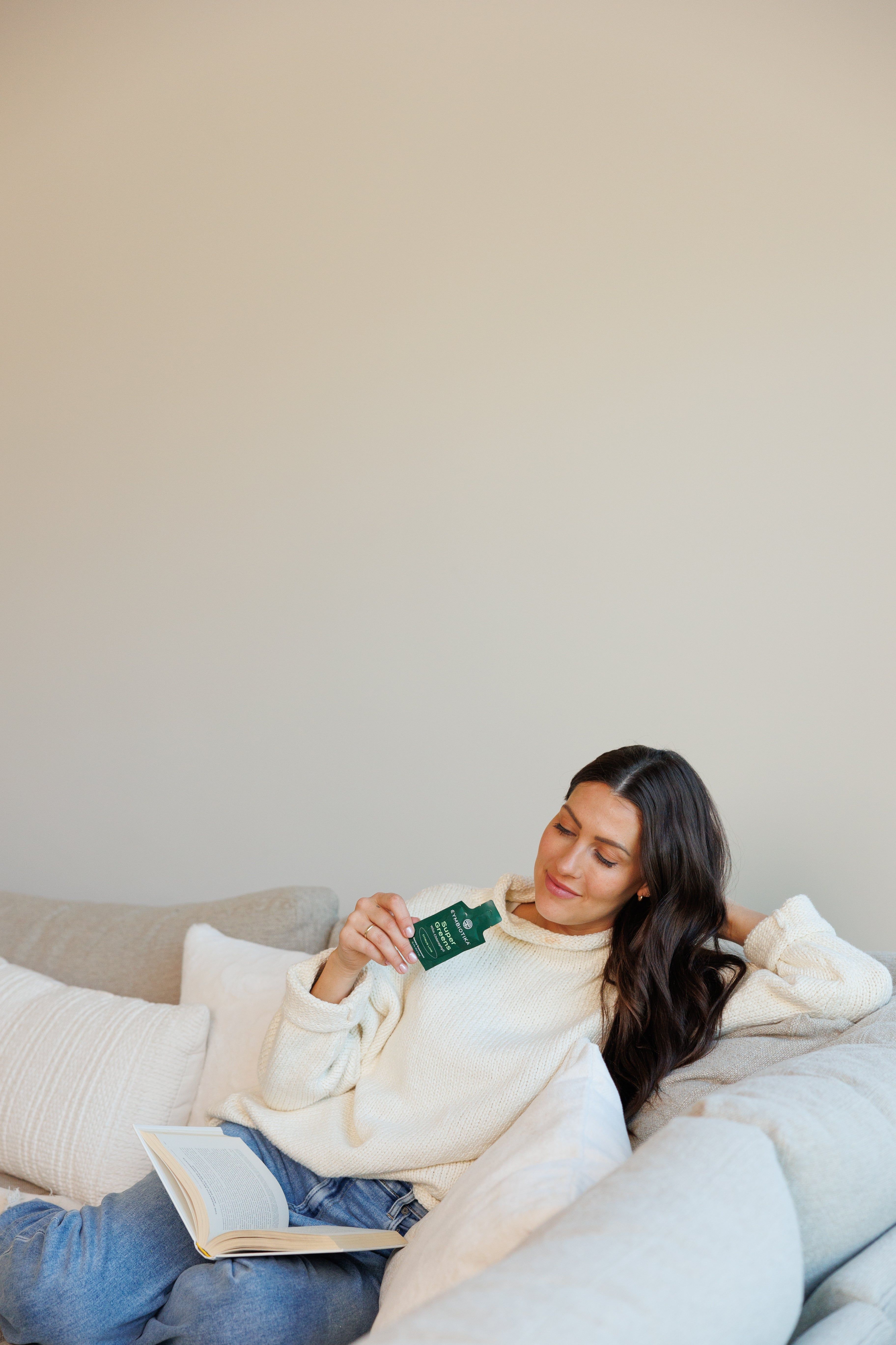 Woman in a cream sweater relaxes on a sofa with an open book and holds a green Cymbiotika supplement packet.