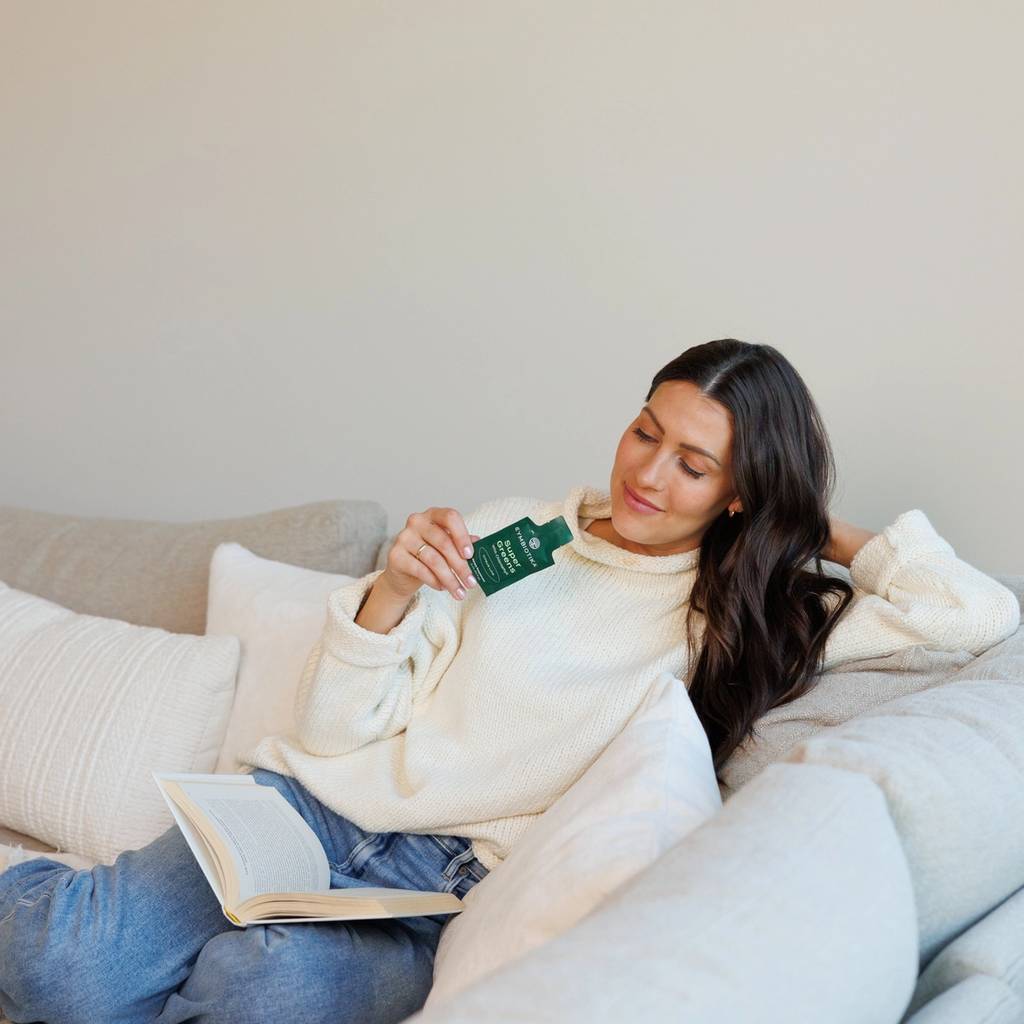 Woman in a cream sweater relaxes on a sofa with an open book and holds a green Cymbiotika supplement packet.