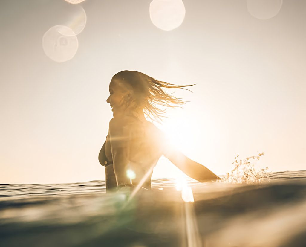 Woman in ocean water at sunset, extending one arm through the surface.
