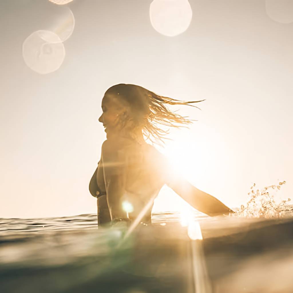 Woman in ocean water at sunset, extending one arm through the surface.
