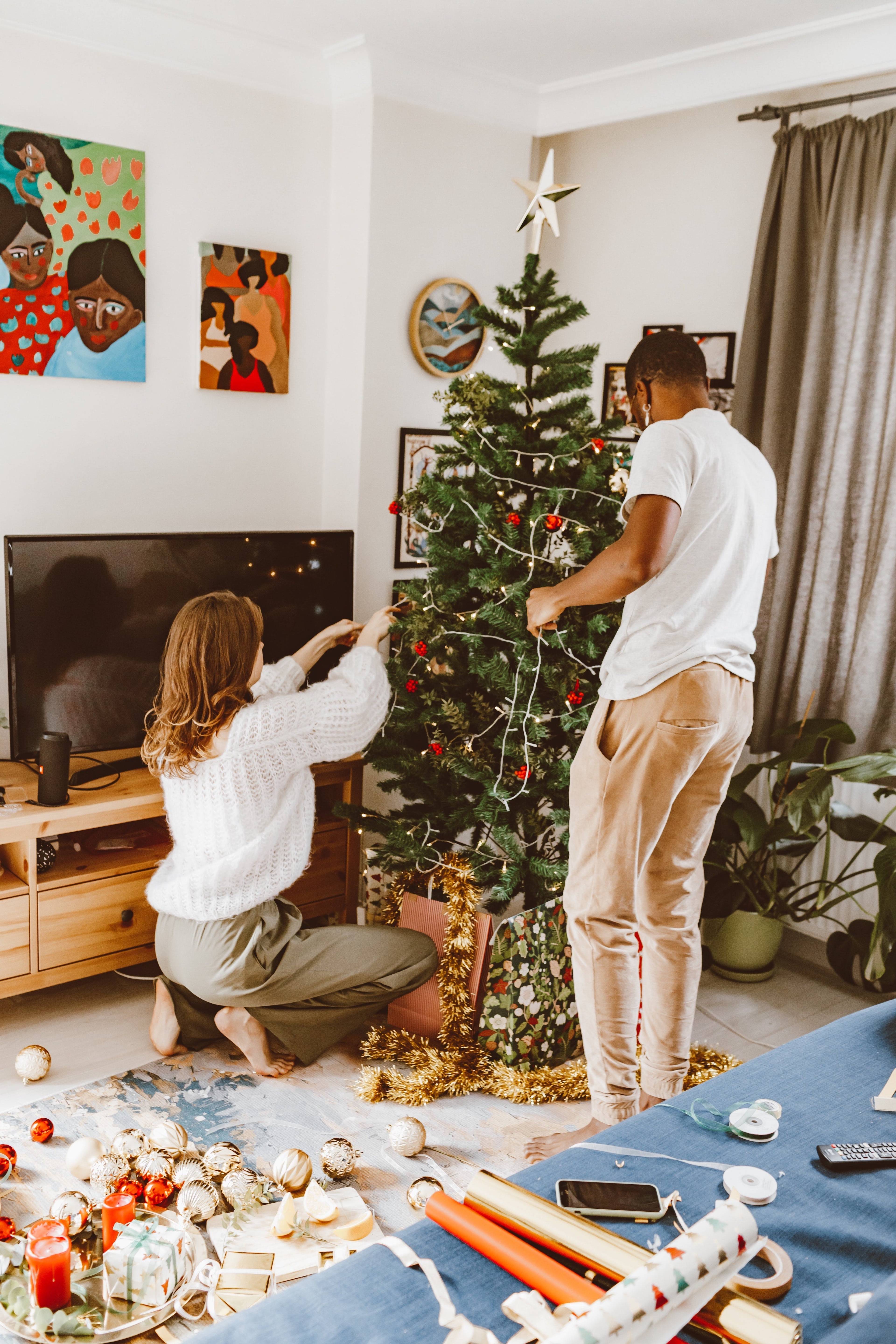 Two people decorate a Christmas tree beside wrapped gifts and holiday craft supplies in a living room.