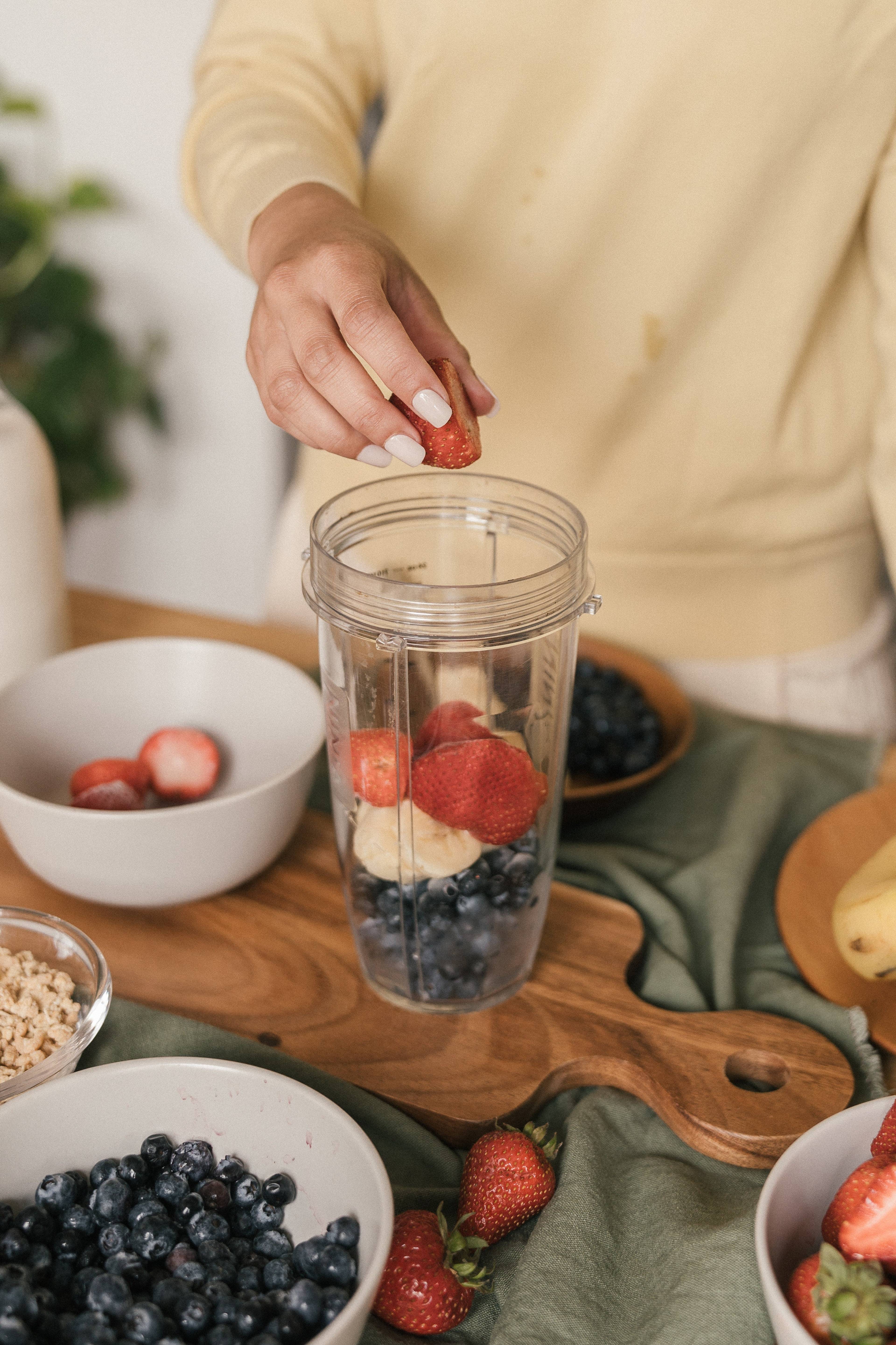 Person adding a strawberry to a blender cup with blueberries and banana on a table.