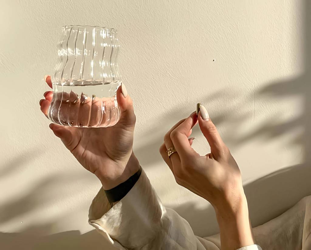 Hands holding a ribbed transparent vessel of water and a small oblong object in warm sunlight.