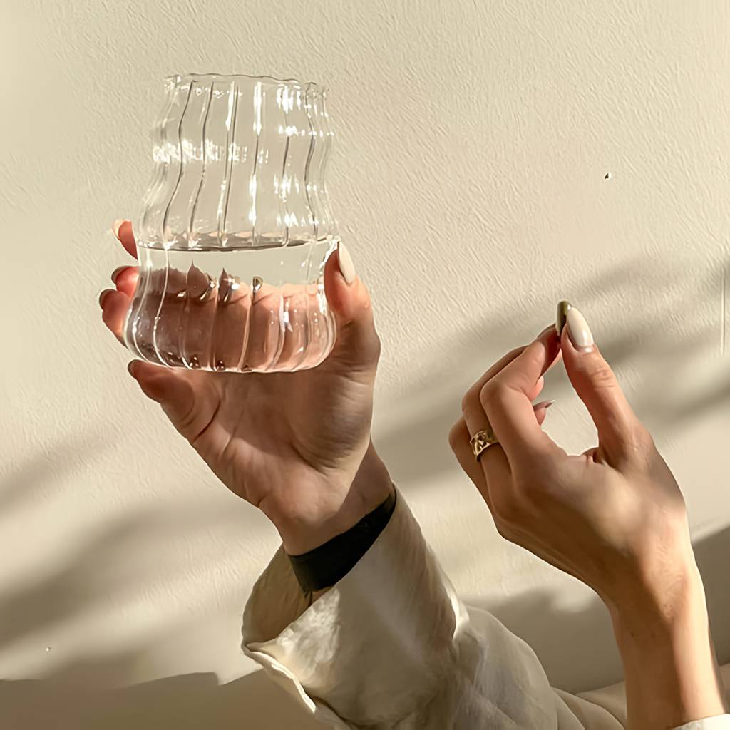 Hands holding a ribbed transparent vessel of water and a small oblong object in warm sunlight.