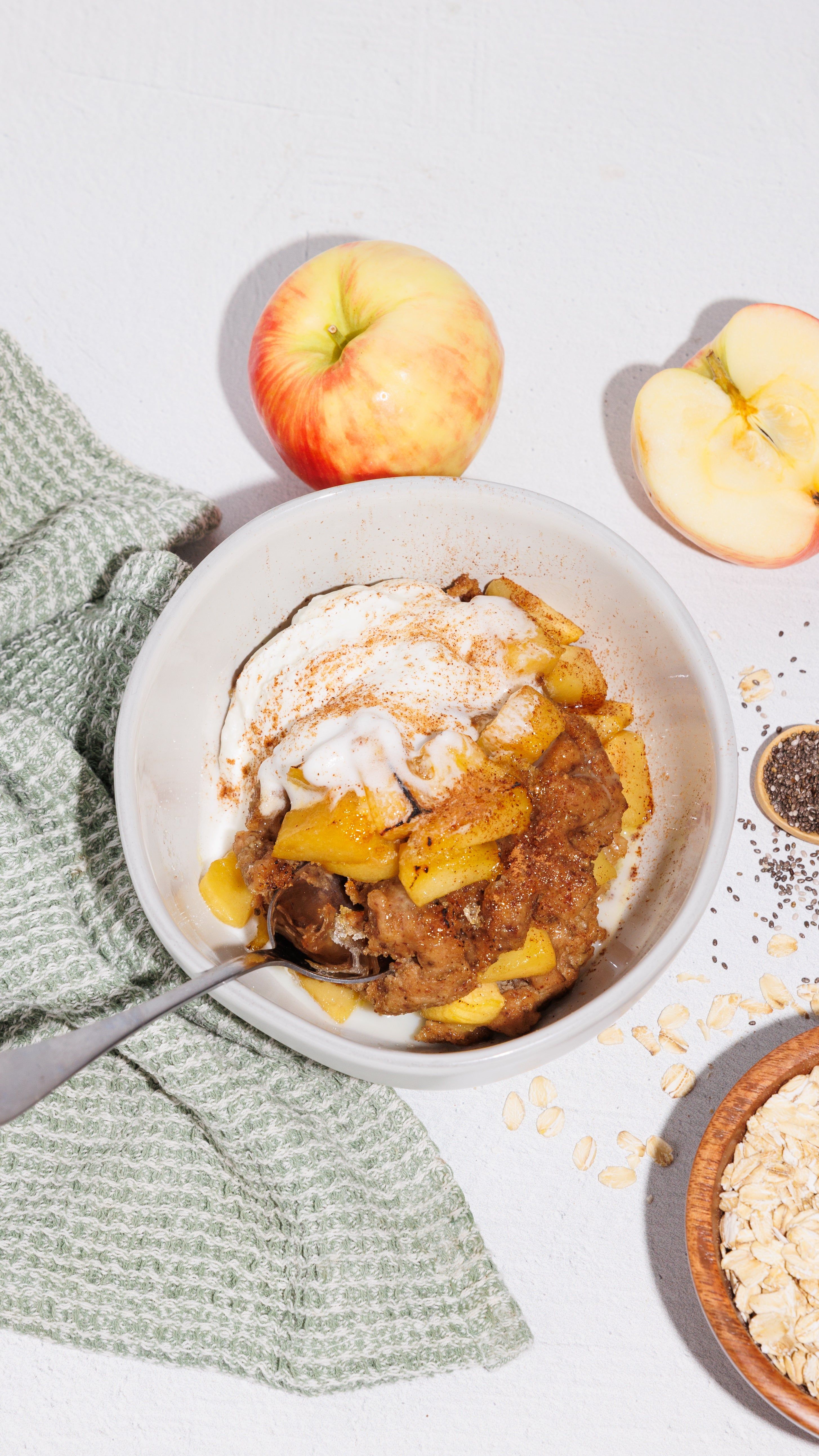Bowl of apple crumble topped with cream, surrounded by fresh apples, oats, chia seeds, and a green cloth.