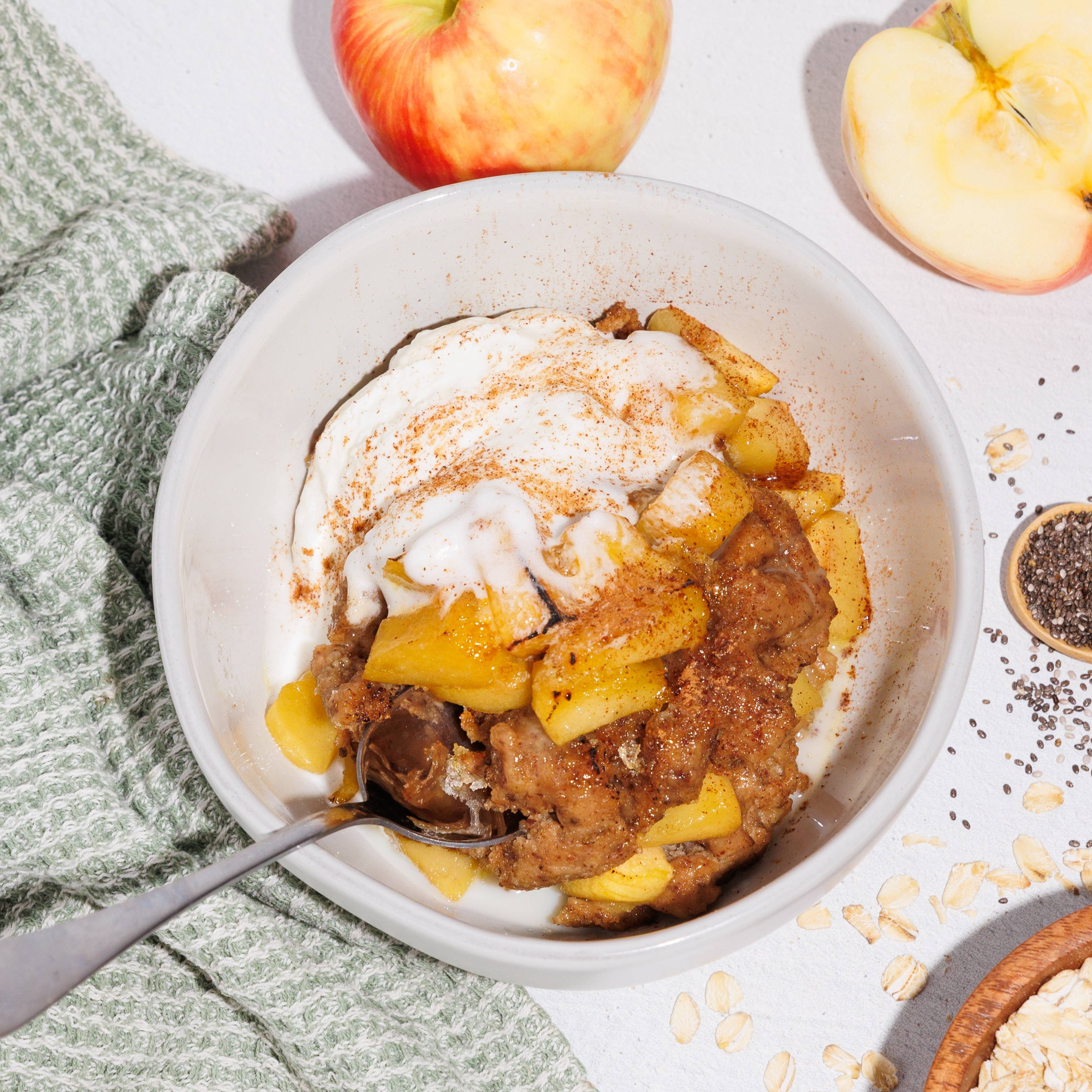 Bowl of apple crumble topped with cream, surrounded by fresh apples, oats, chia seeds, and a green cloth.
