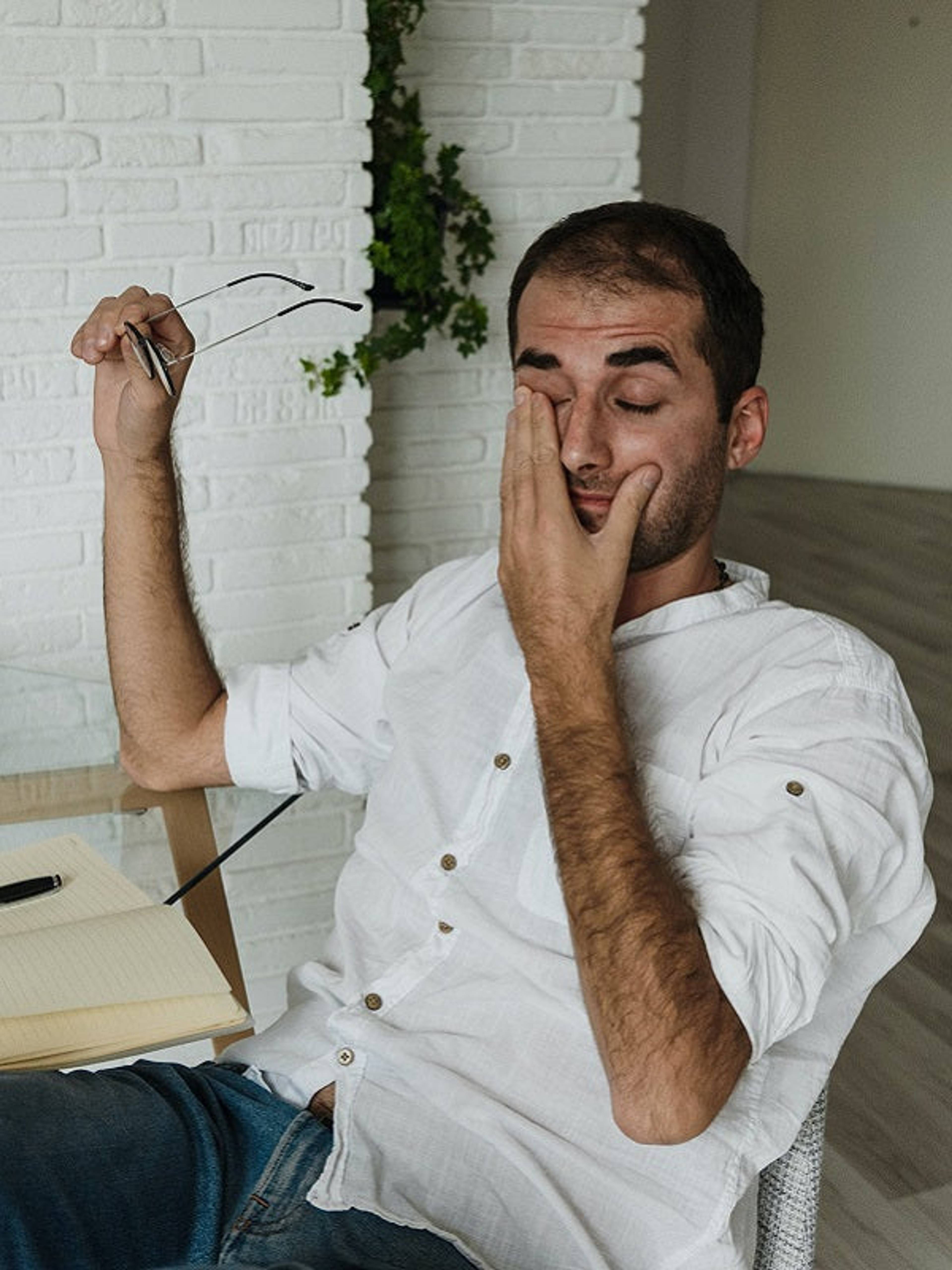 Tired man at a desk rubs his eye while holding eyeglasses.