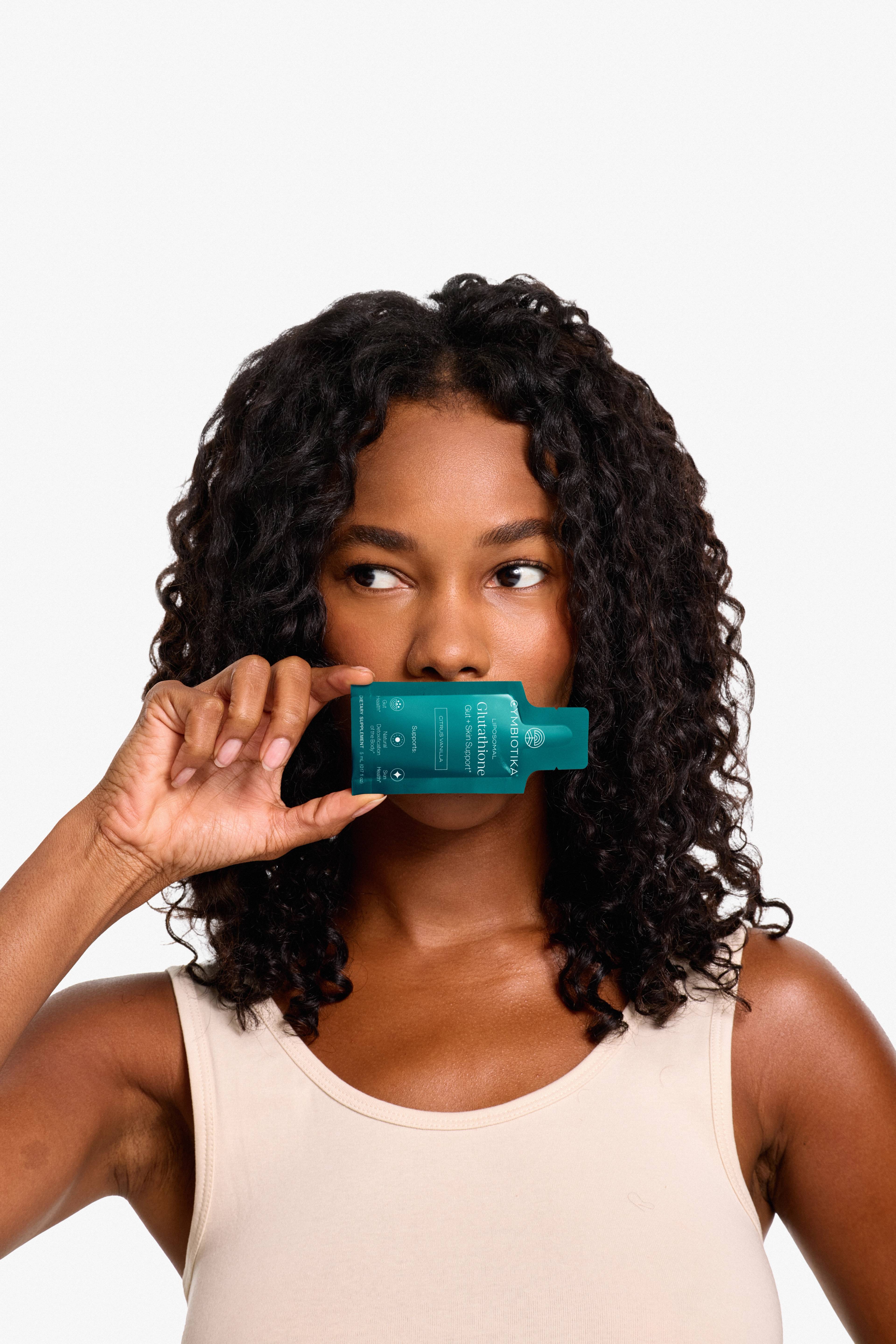 Woman holding a Cymbiotika Liposomal Glutathione packet beneath her nose against a white background.
