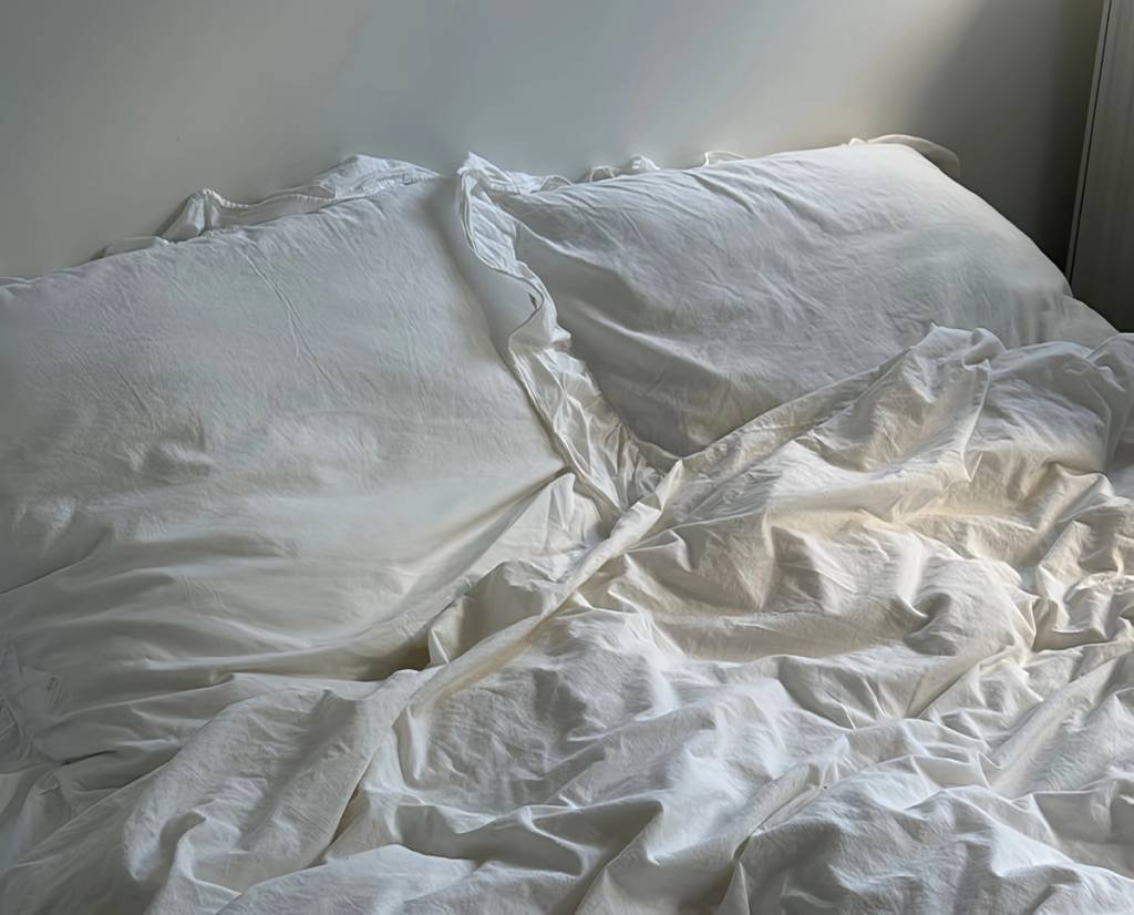 An unmade bed with rumpled white sheets and two pillows in soft daylight.