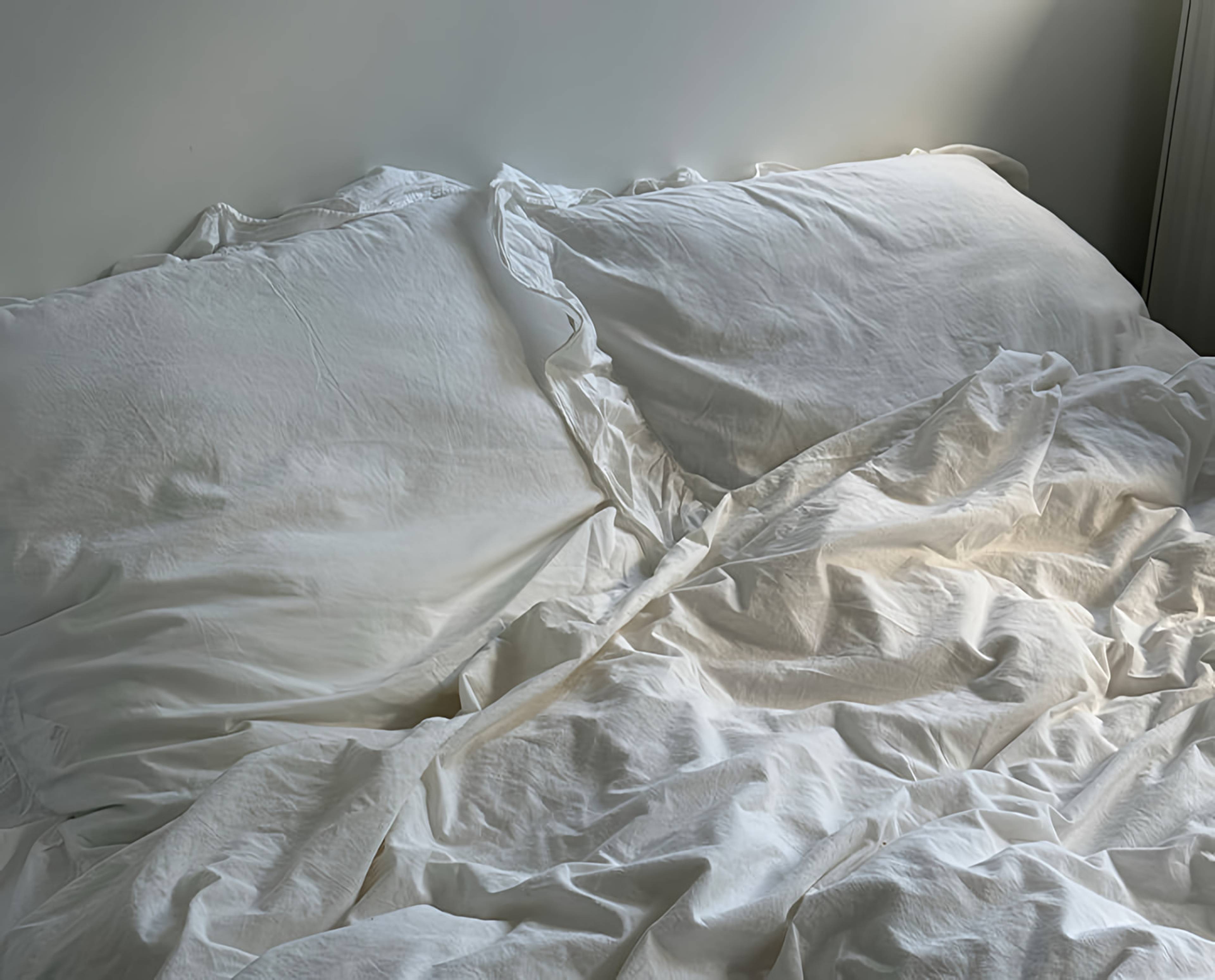 An unmade bed with rumpled white sheets and two pillows in soft daylight.