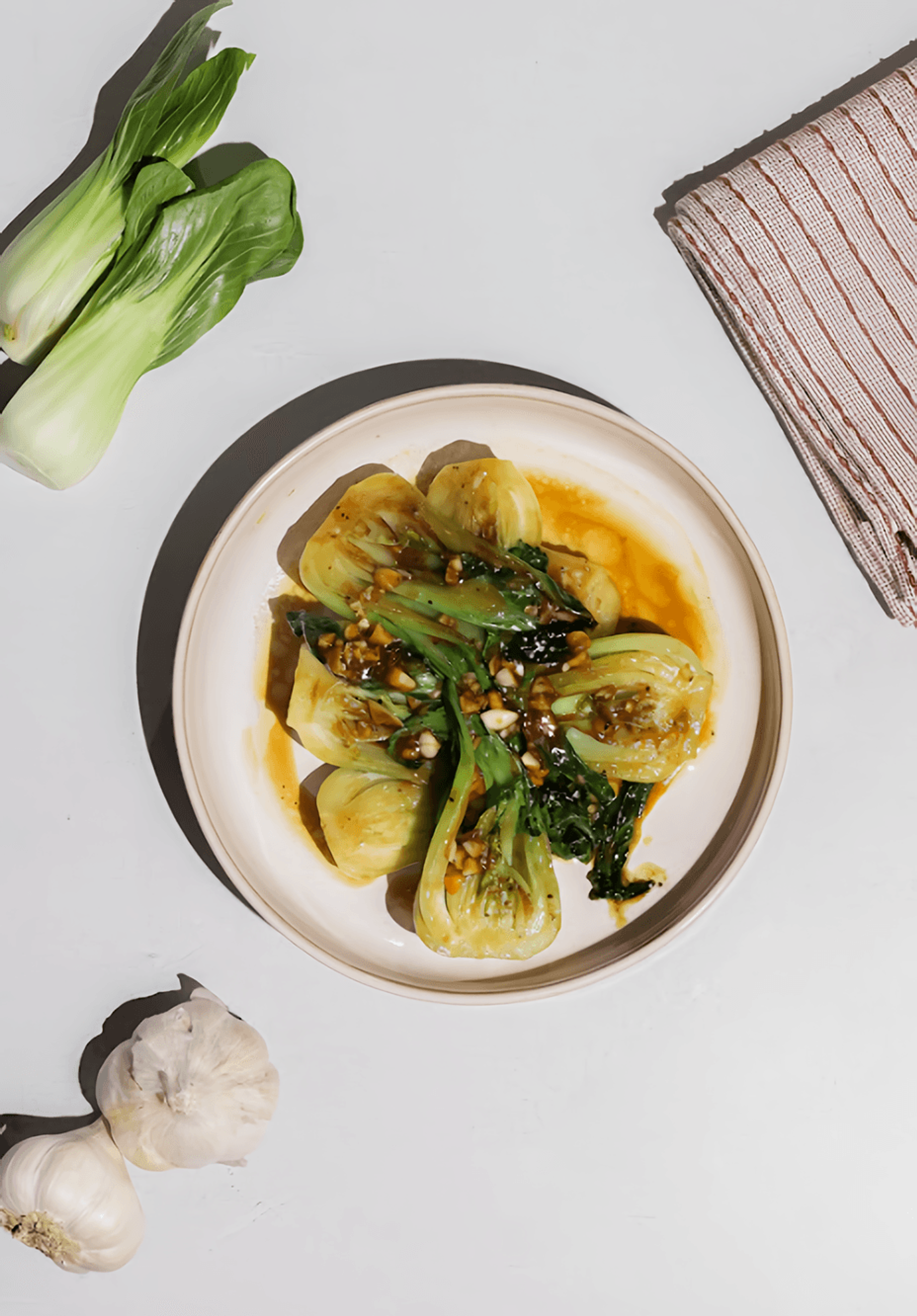 Overhead view of sauced bok choy in a bowl with fresh bok choy, garlic bulbs, and a striped kitchen towel.