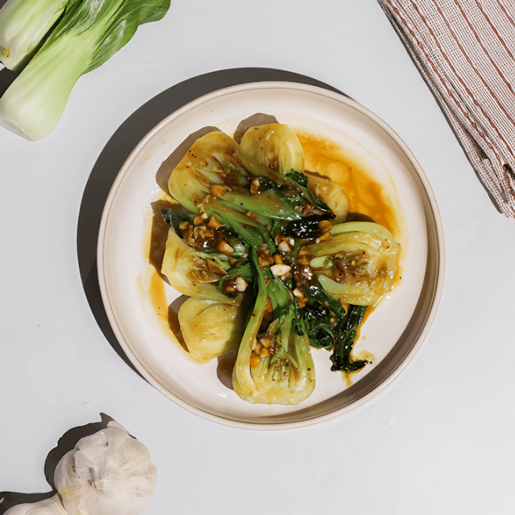 Overhead view of sauced bok choy in a bowl with fresh bok choy, garlic bulbs, and a striped kitchen towel.