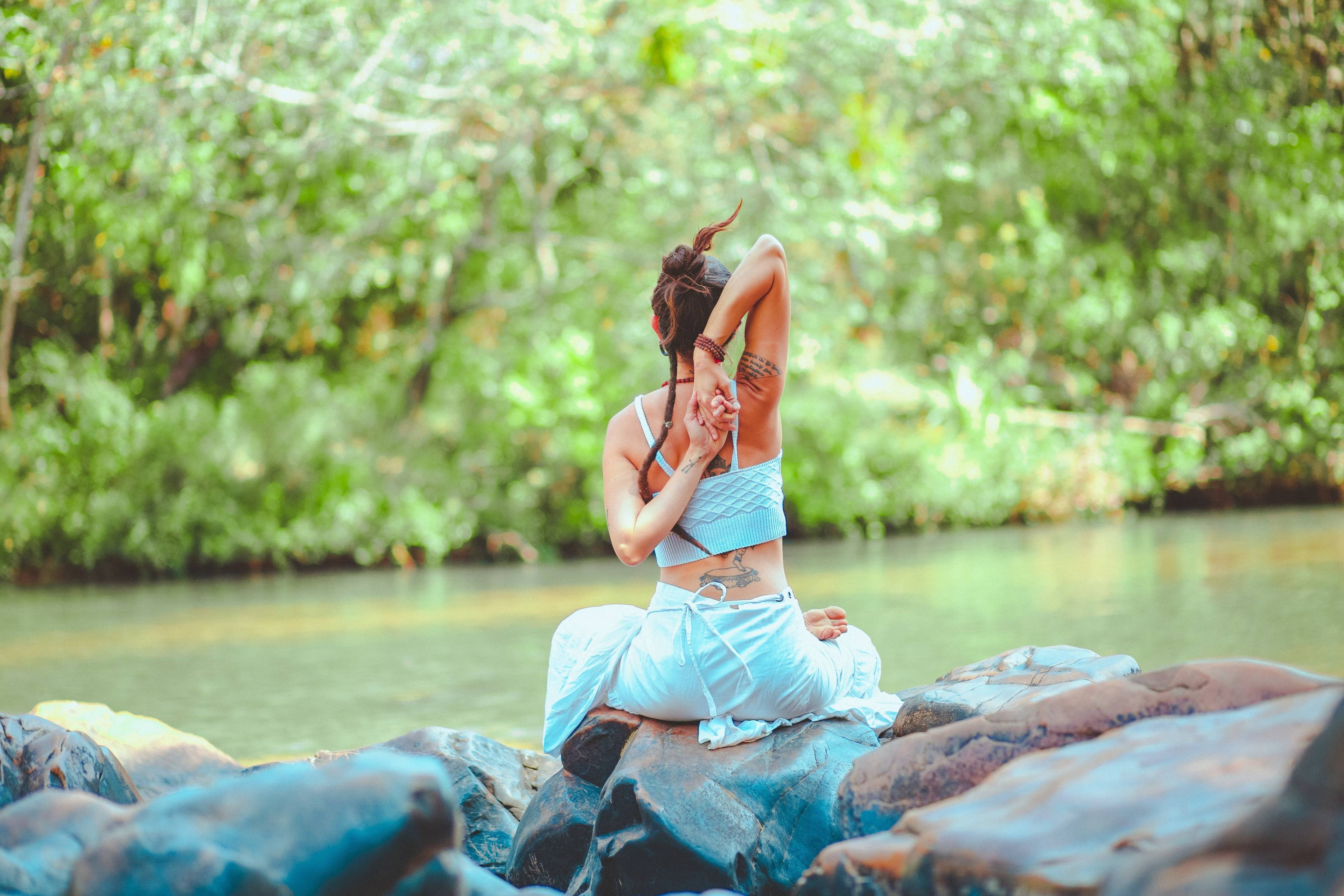 Woman seated cross-legged on riverside rocks, stretching with her arms clasped behind her back.