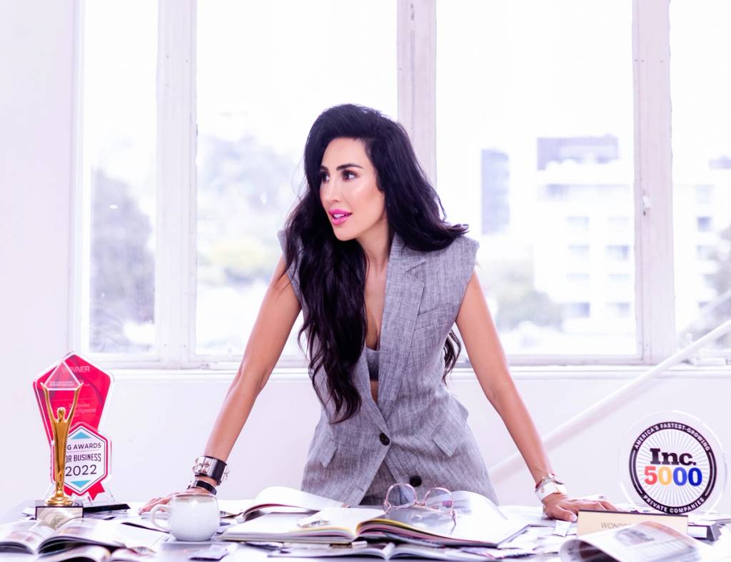 Woman in a gray vest leans over a desk with papers, a cup, and business awards.