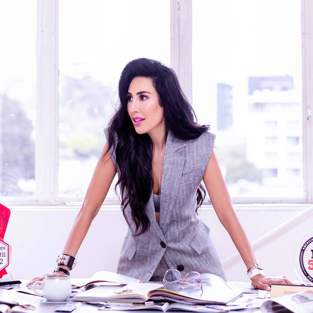 Woman in a gray vest leans over a desk with papers, a cup, and business awards.
