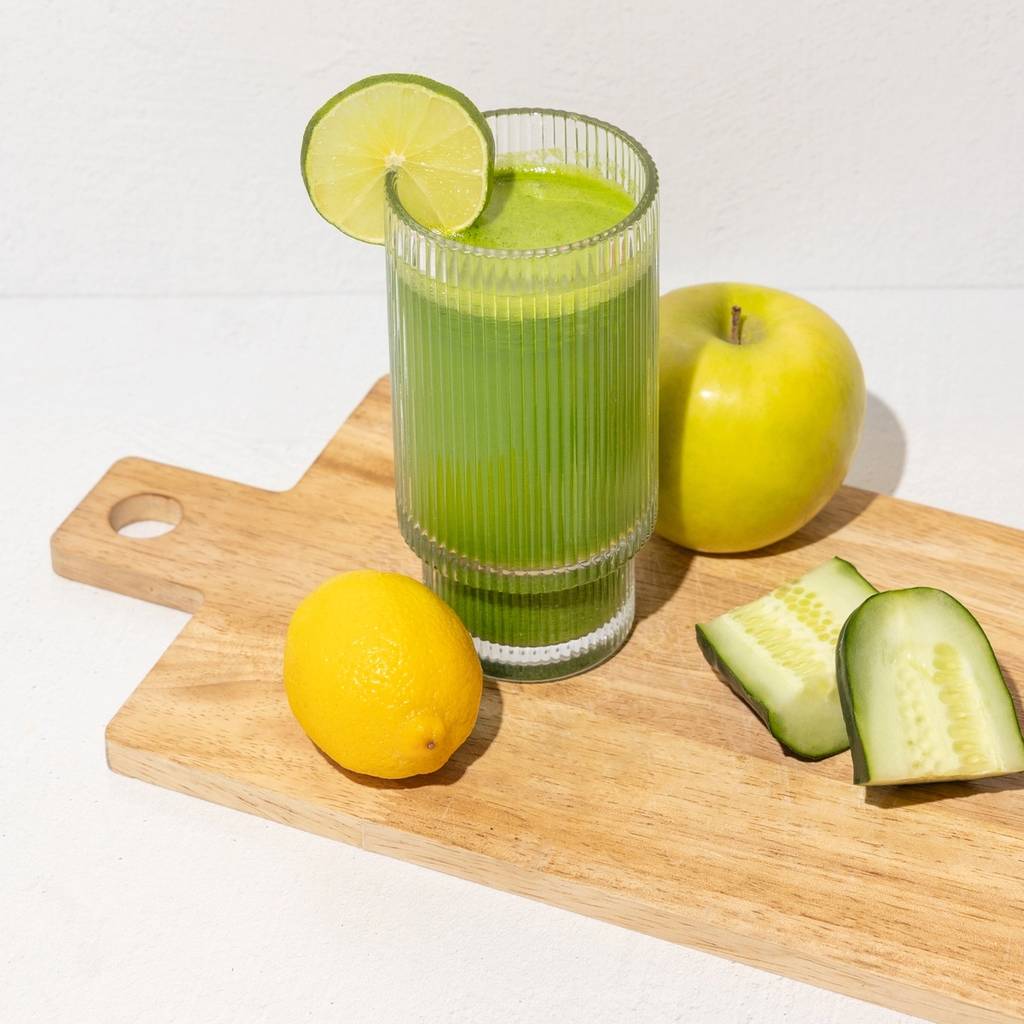 Glass of green juice with a lime slice, apple, lemon, and cucumber on a wooden cutting board.
