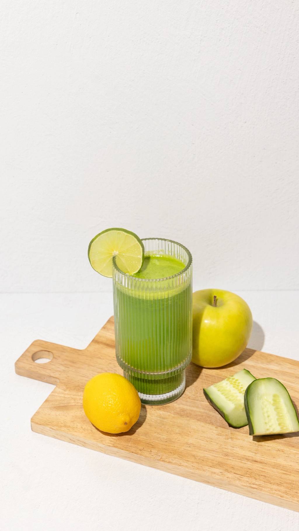 Glass of green juice with a lime slice, apple, lemon, and cucumber on a wooden cutting board.