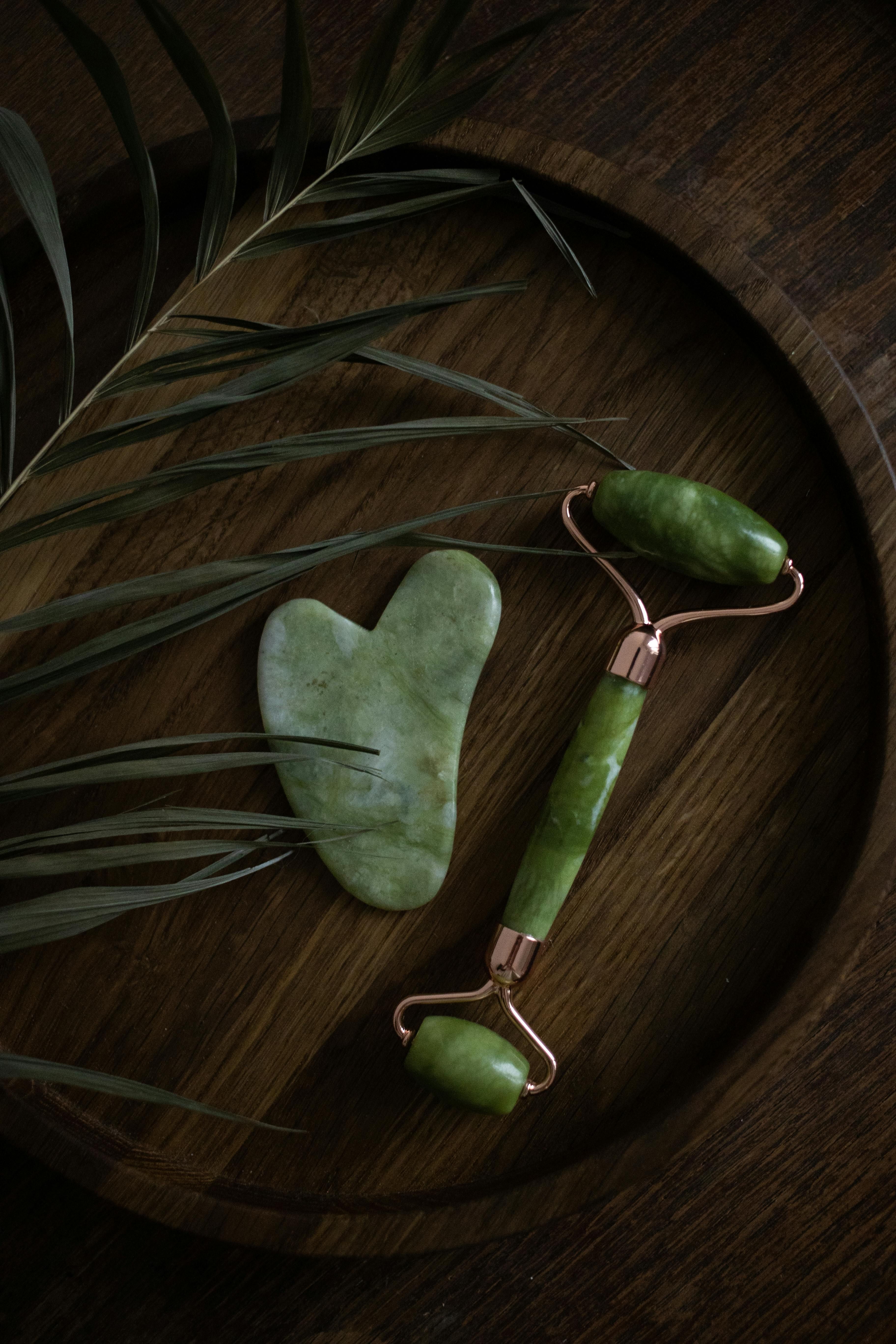 Green stone gua sha tool and facial roller on a round wooden tray with palm leaves.