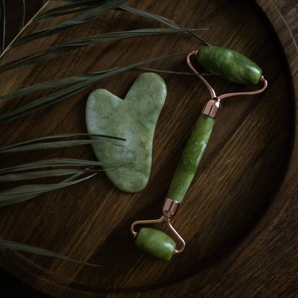 Green stone gua sha tool and facial roller on a round wooden tray with palm leaves.
