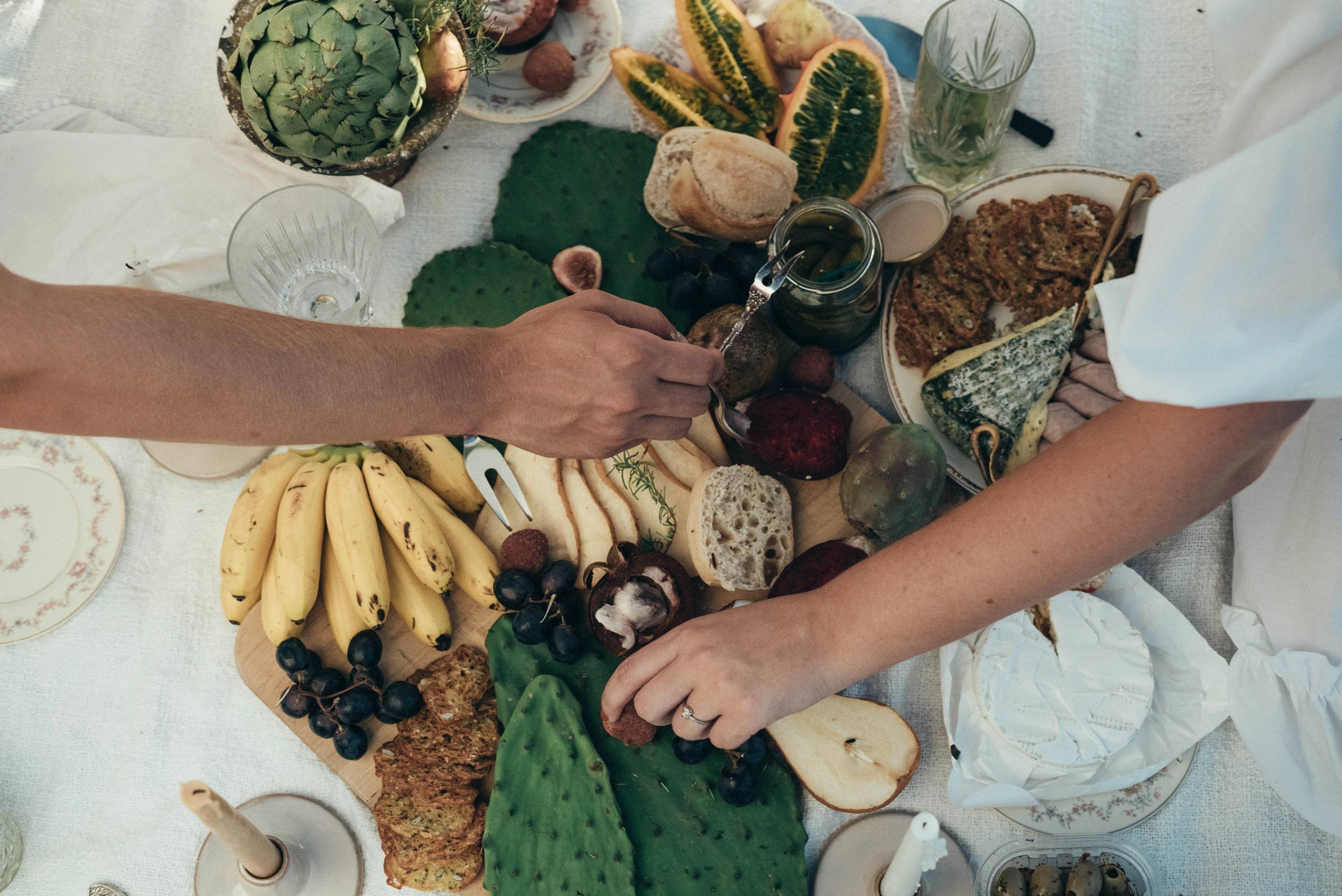 Overhead view of two hands reaching across a picnic spread with bananas, grapes, bread, cheese, artichokes, and other foods.