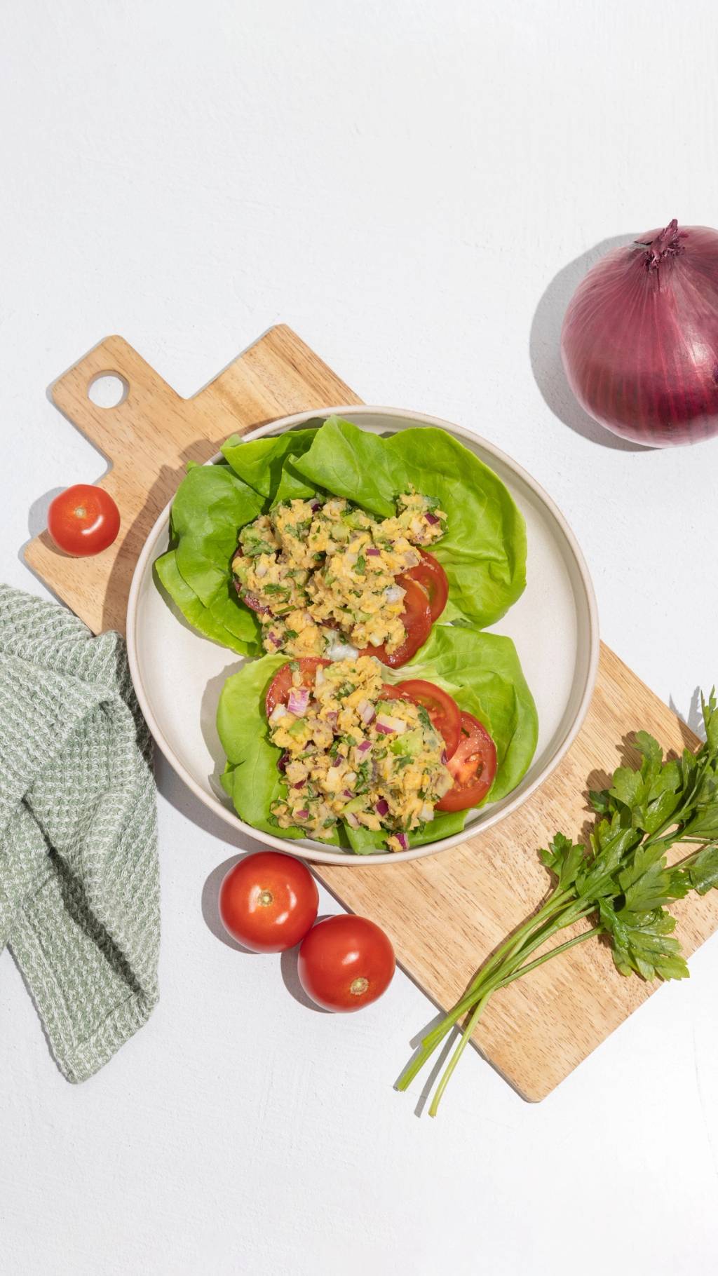 Two lettuce cups filled with chopped salad and tomato slices on a cutting board, with whole tomatoes, a red onion, and parsley nearby.