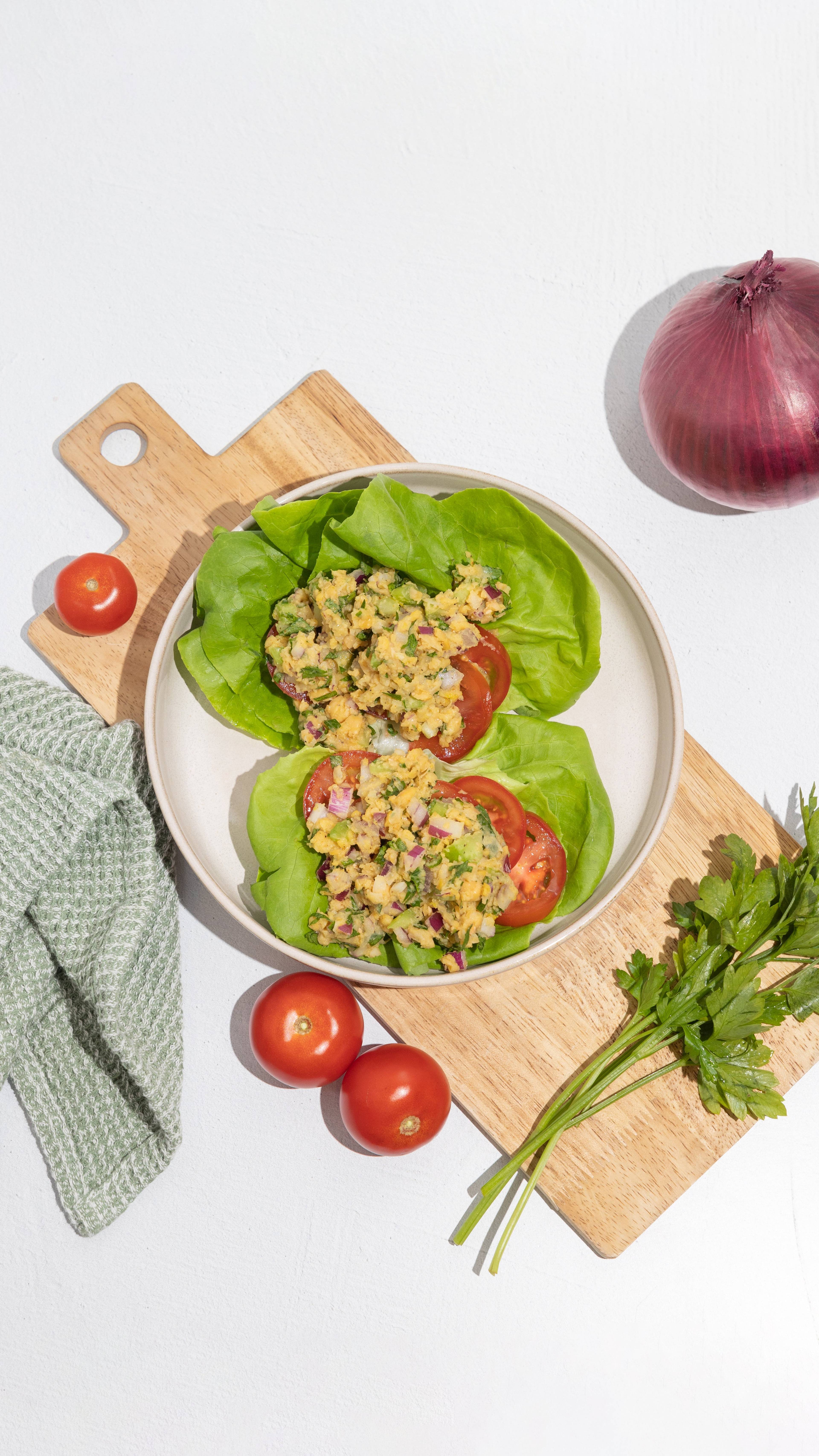Two lettuce cups filled with chopped salad and tomato slices on a cutting board, with whole tomatoes, a red onion, and parsley nearby.