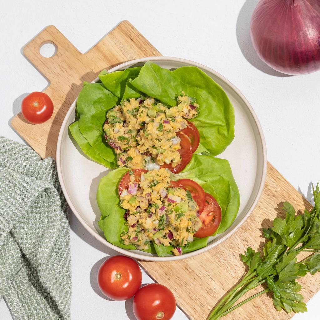 Two lettuce cups filled with chopped salad and tomato slices on a cutting board, with whole tomatoes, a red onion, and parsley nearby.