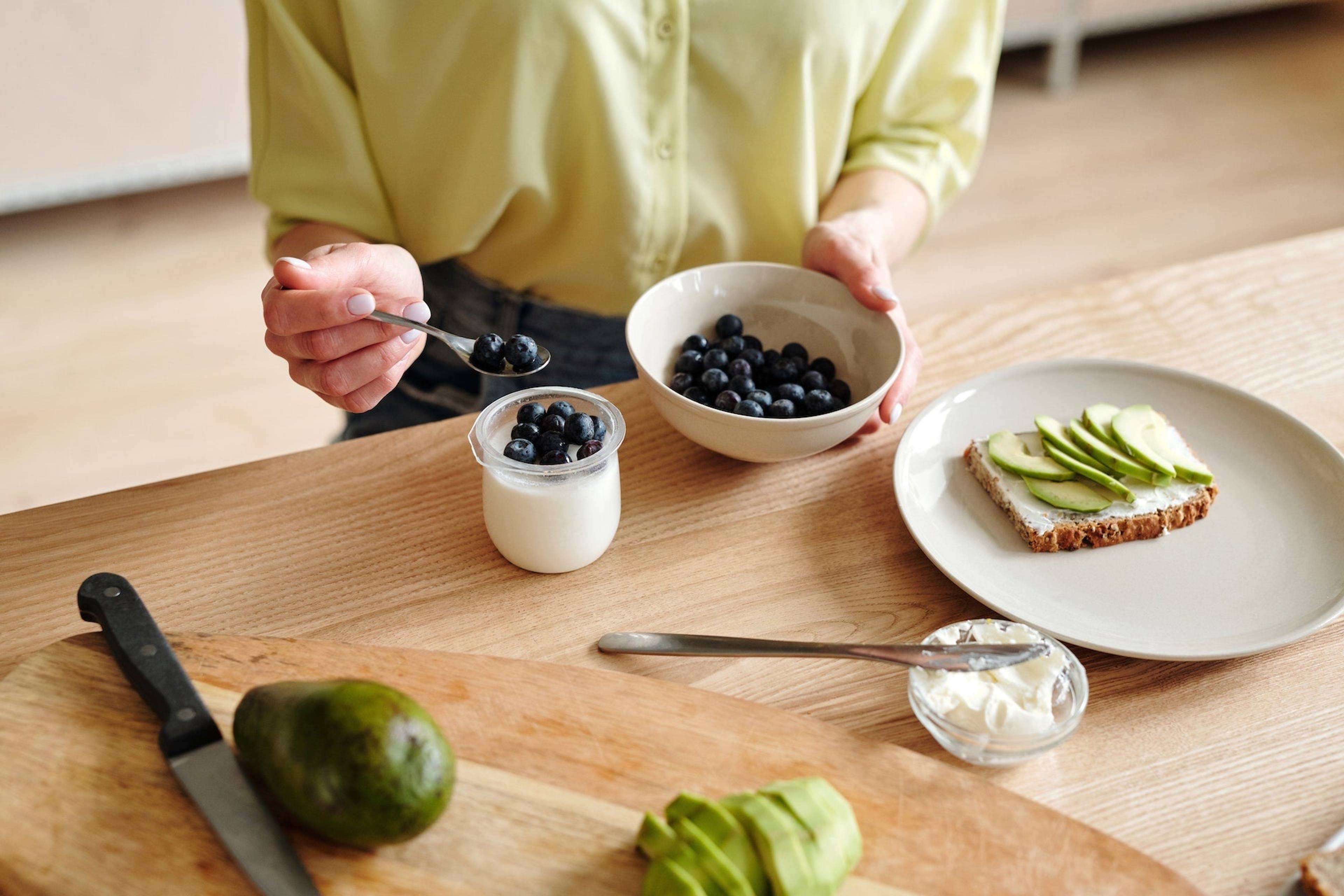 A person holds a spoonful of blueberries over a cup of yogurt, with avocado toast and a bowl of blueberries on a wooden table.