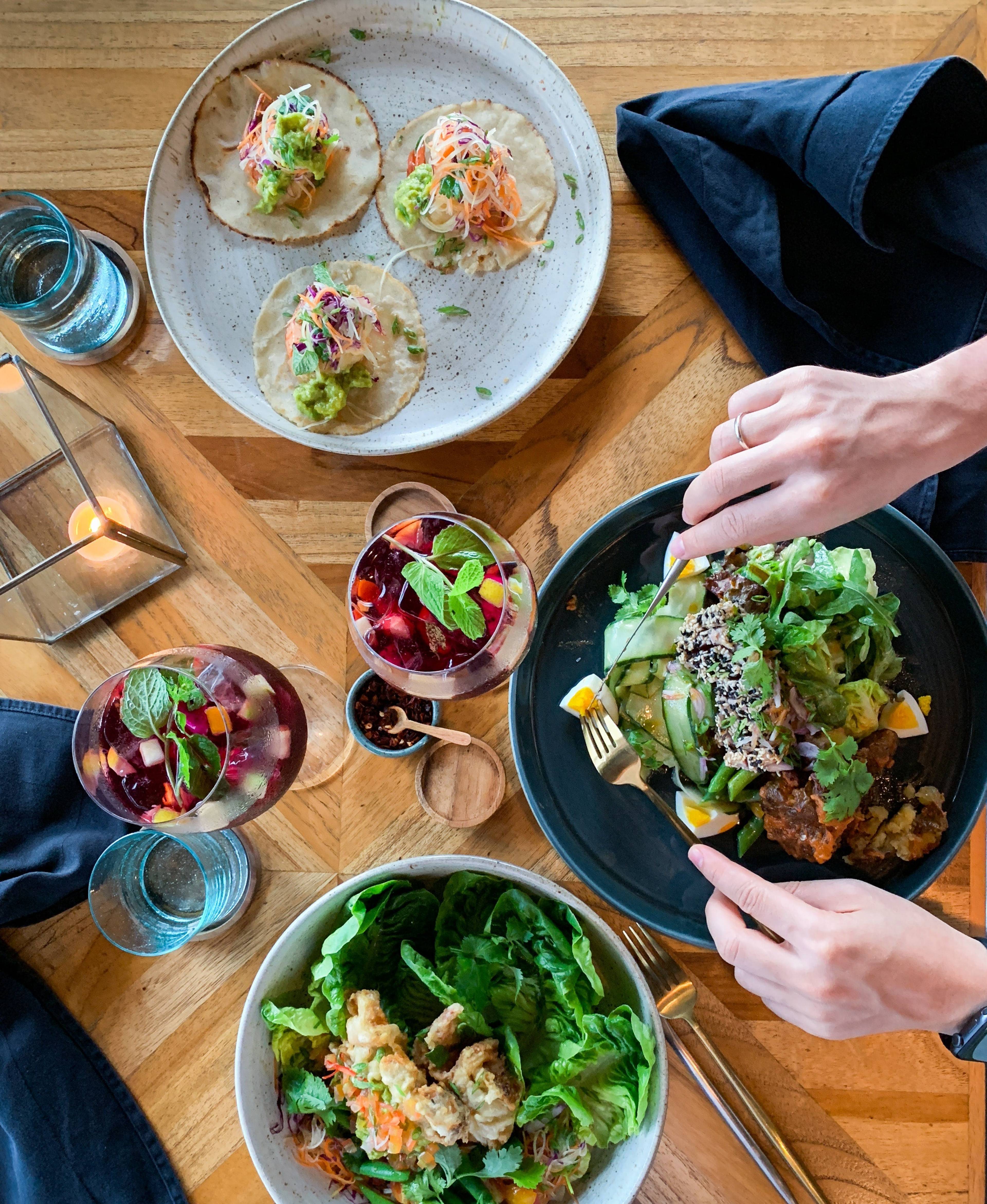 Overhead view of tacos, salads, fruit drinks, and water on a wooden table as a person eats.