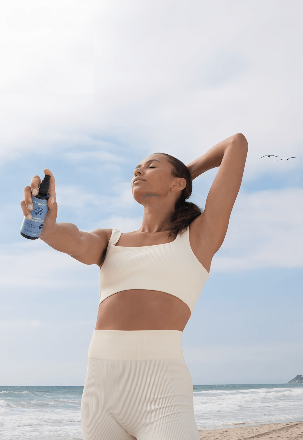 Woman in cream activewear holds a blue spray bottle on a beach with her eyes closed.