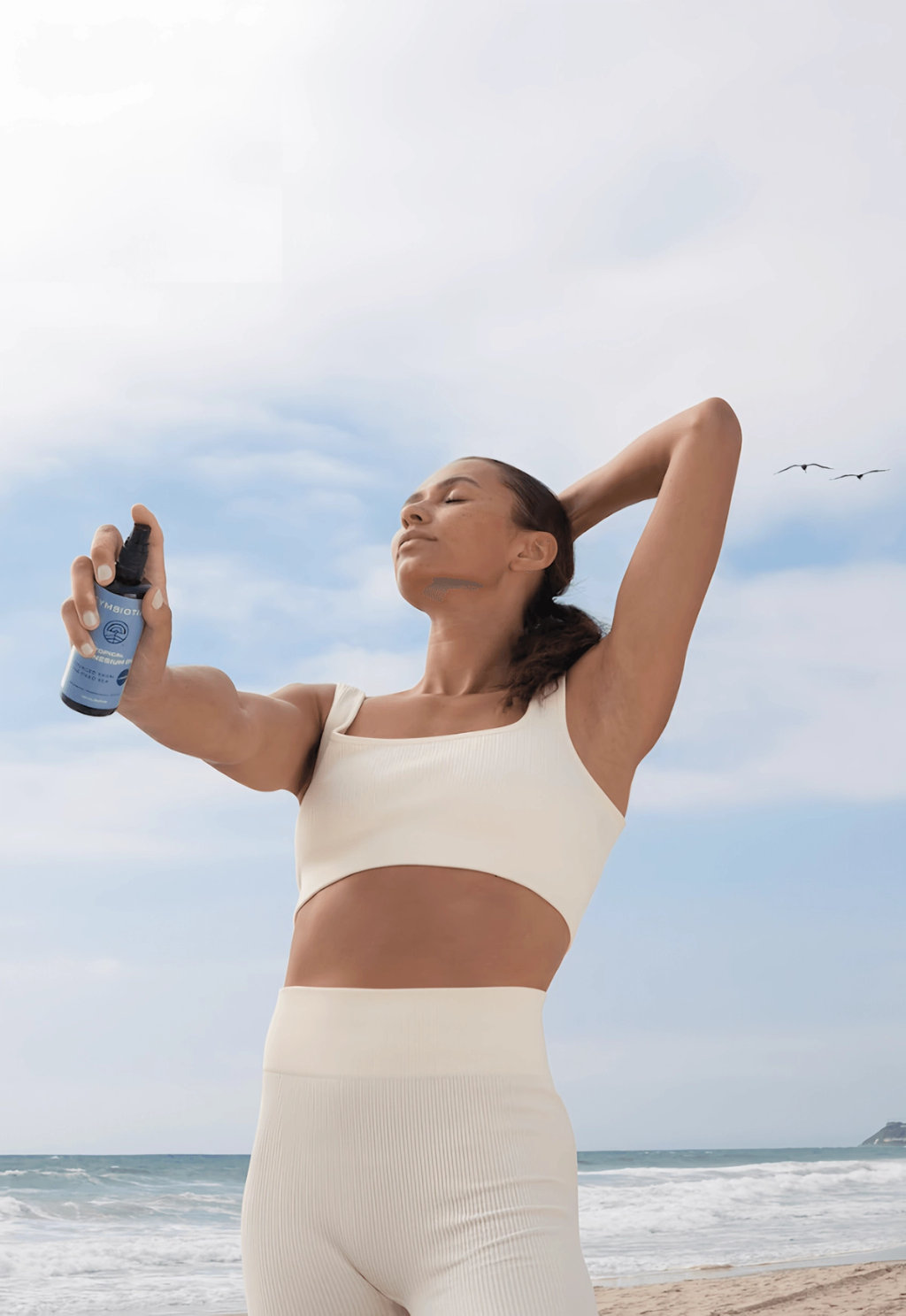 Woman in cream activewear holds a blue spray bottle on a beach with her eyes closed.