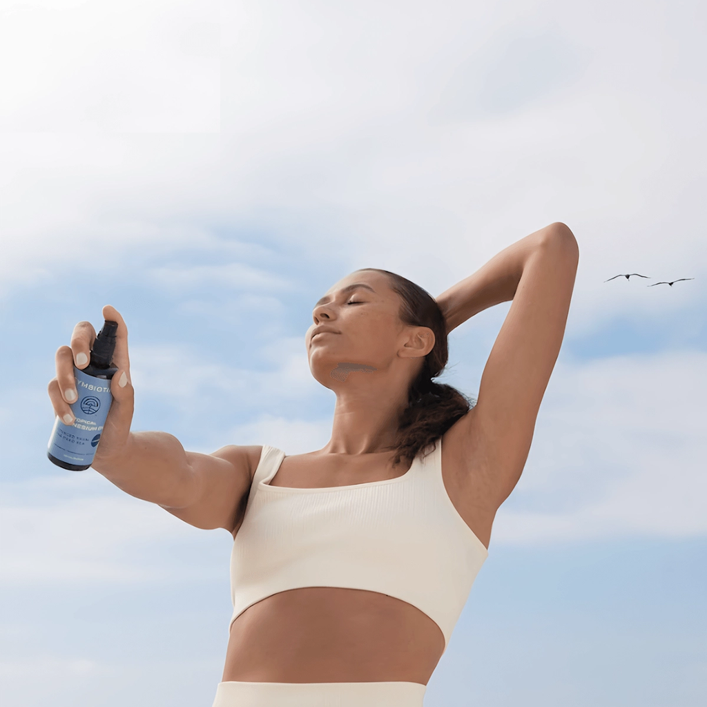 Woman in cream activewear holds a blue spray bottle on a beach with her eyes closed.