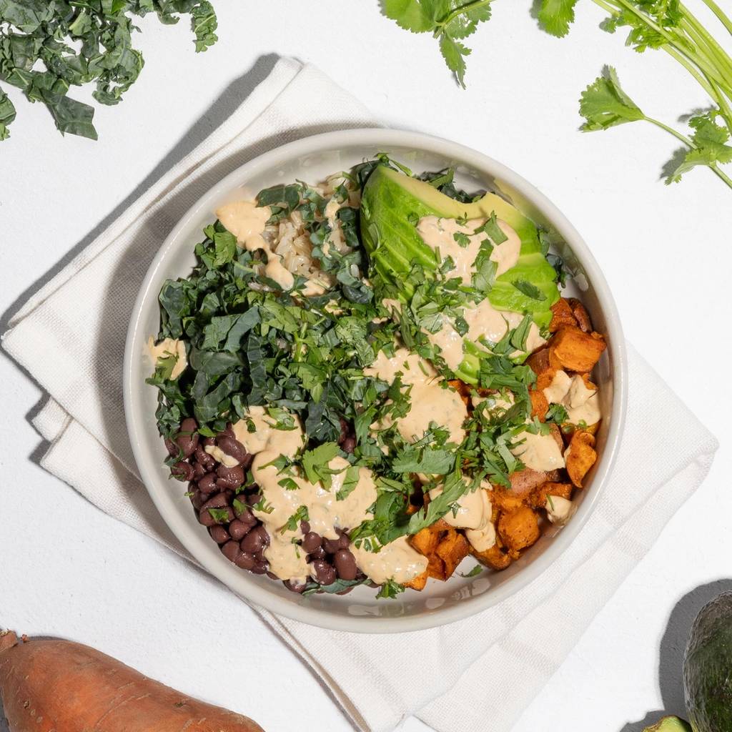 Buddha bowl with sweet potatoes, black beans, kale, avocado, grains, and creamy dressing.