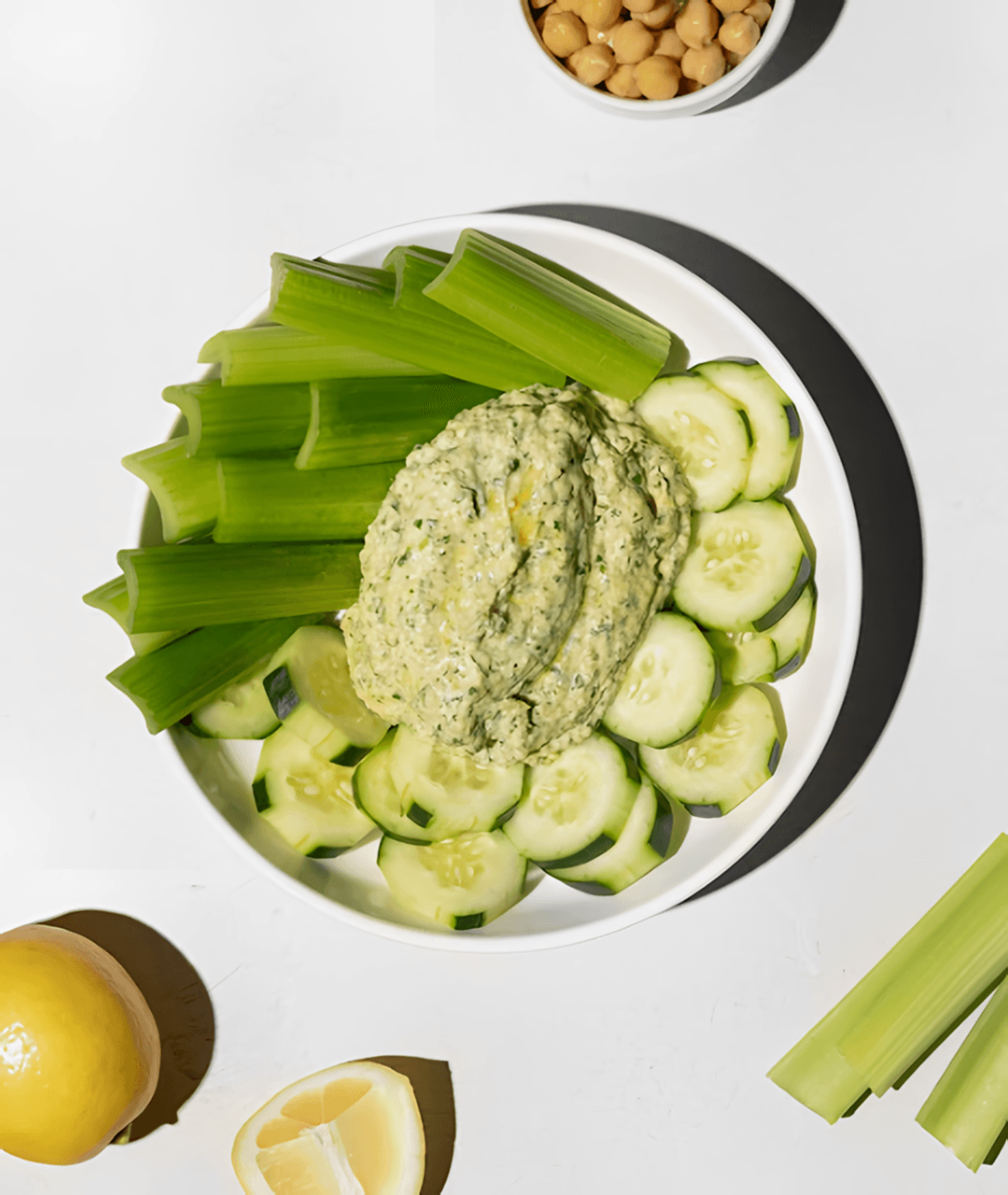 Bowl of green hummus with celery and cucumber slices, with chickpeas and lemon nearby.