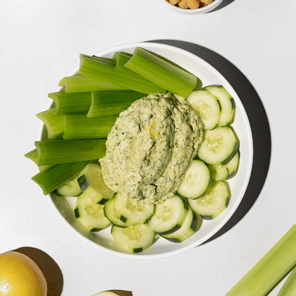 Bowl of green hummus with celery and cucumber slices, with chickpeas and lemon nearby.