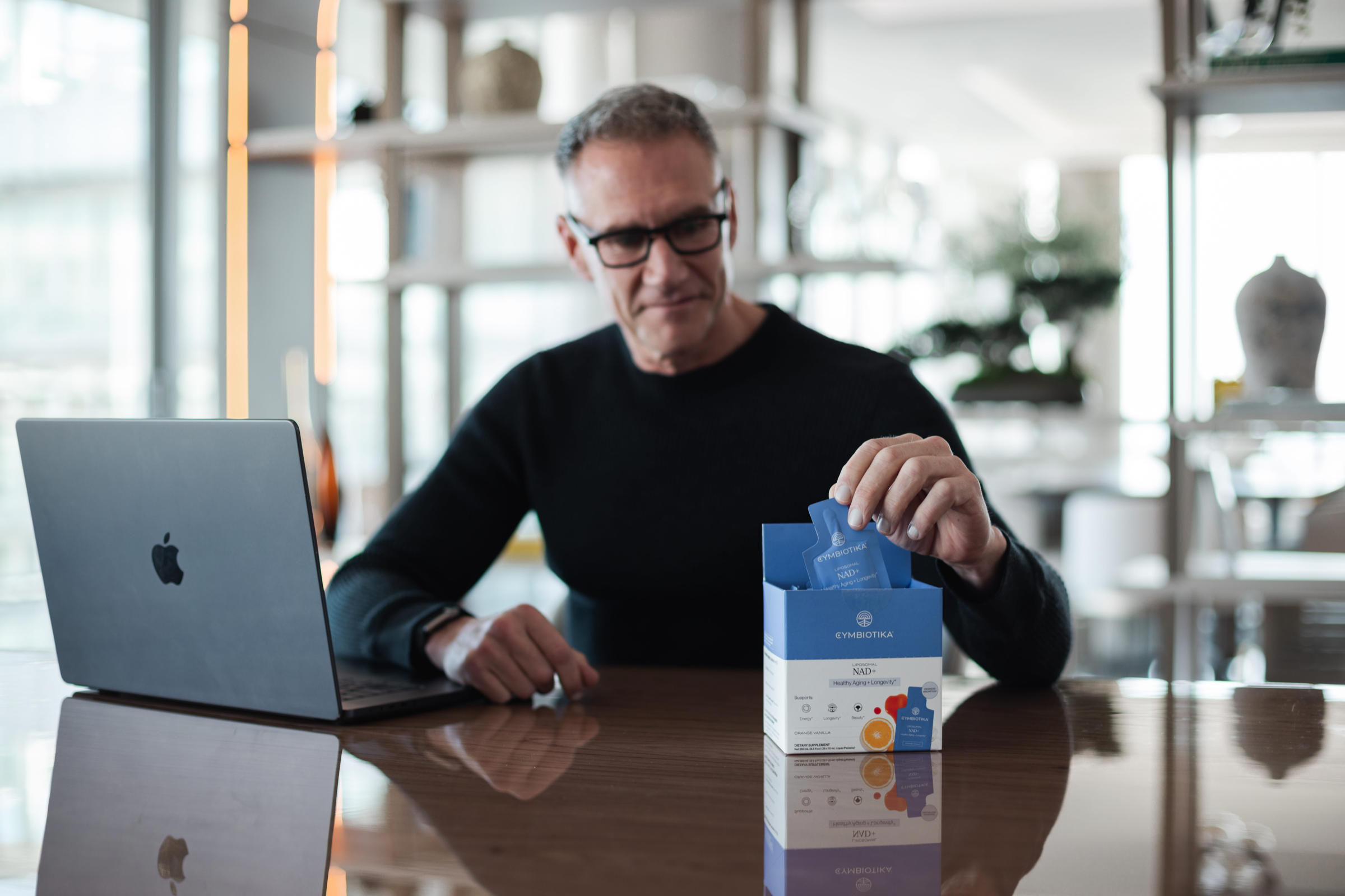 A man at a desk takes a blue sachet from an open Cymbiotika NAD+ box beside a laptop.