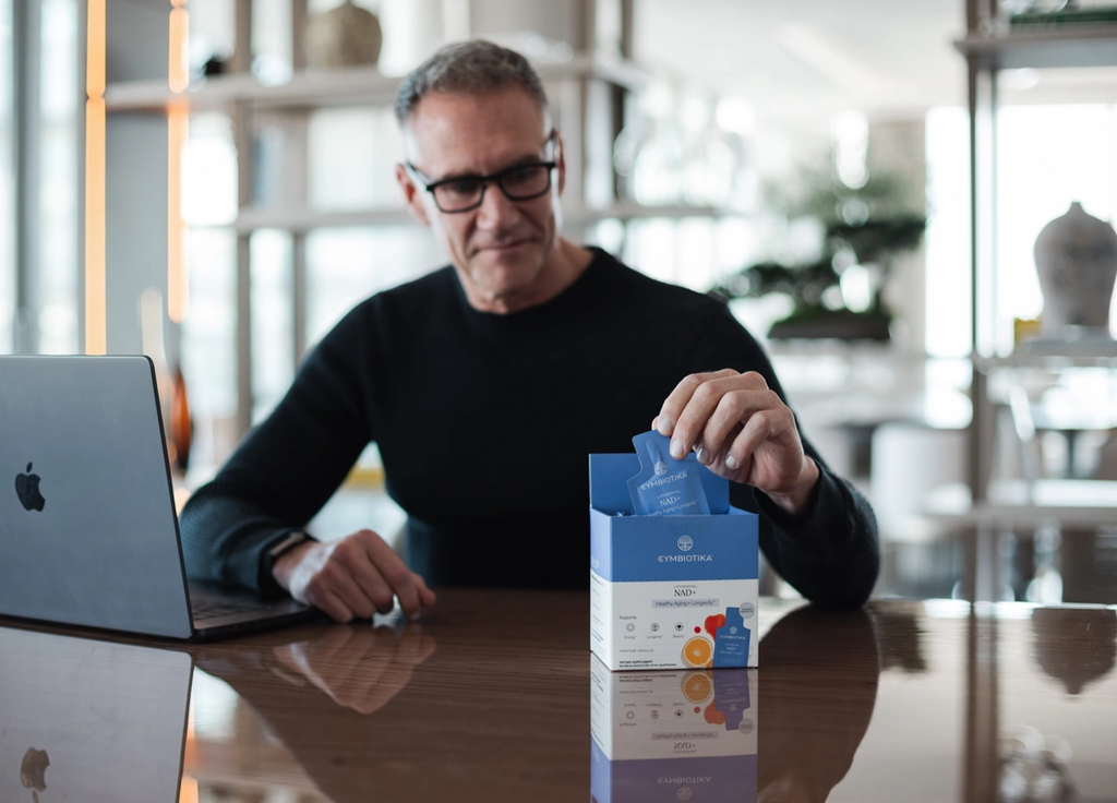 A man at a desk takes a blue sachet from an open Cymbiotika NAD+ box beside a laptop.