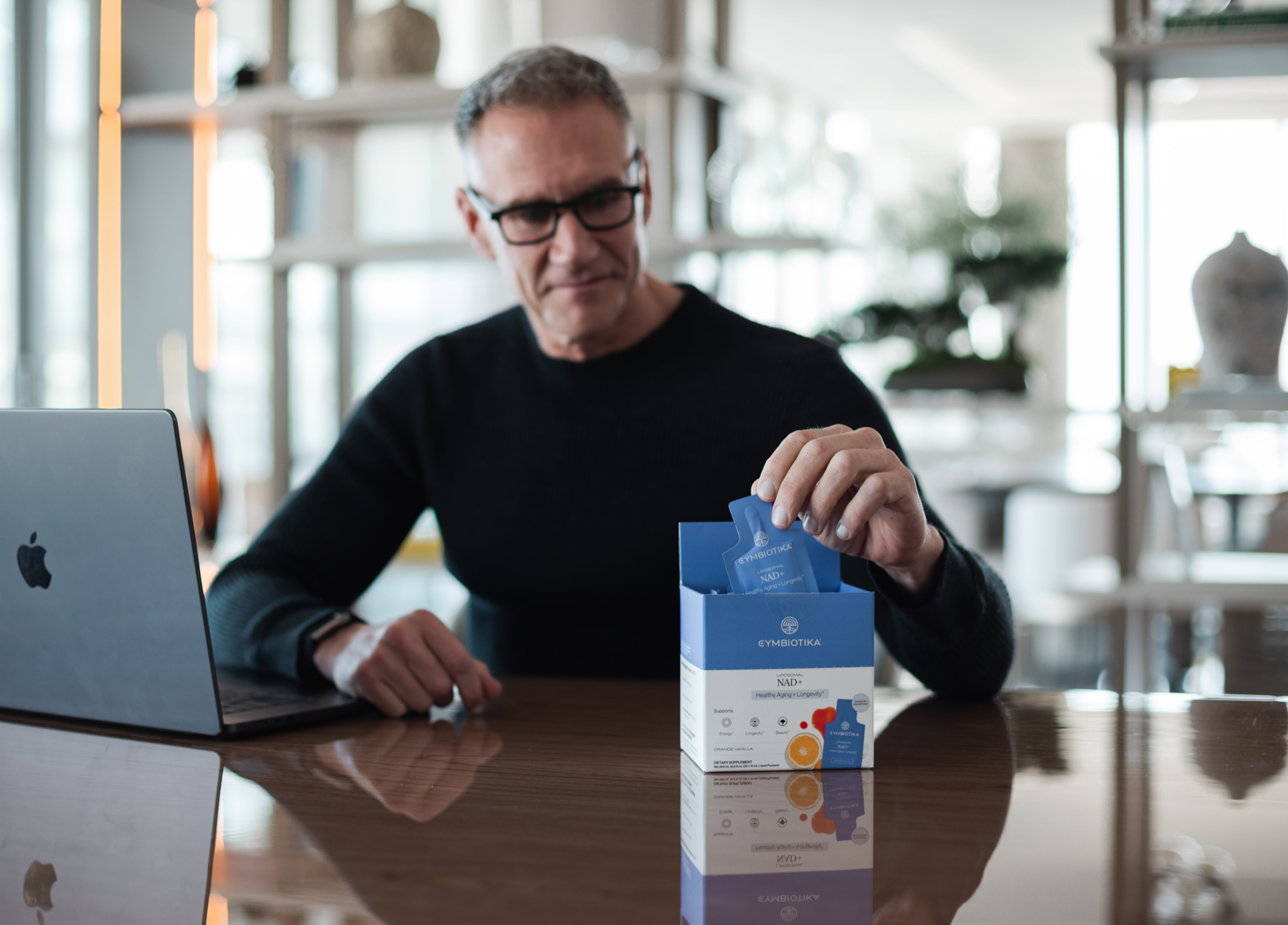 A man at a desk takes a blue sachet from an open Cymbiotika NAD+ box beside a laptop.
