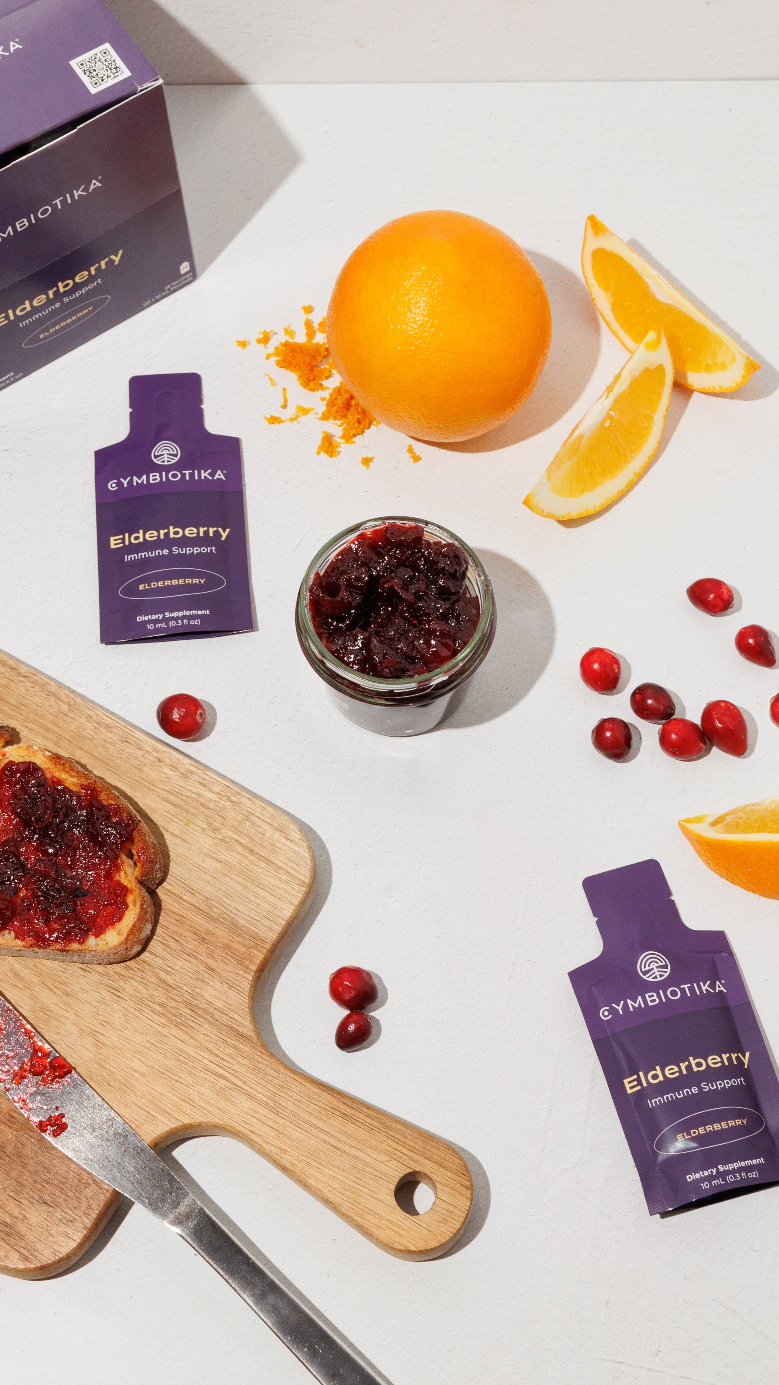 Elderberry supplement packets beside cranberry jam, oranges, cranberries, and toast on a cutting board.