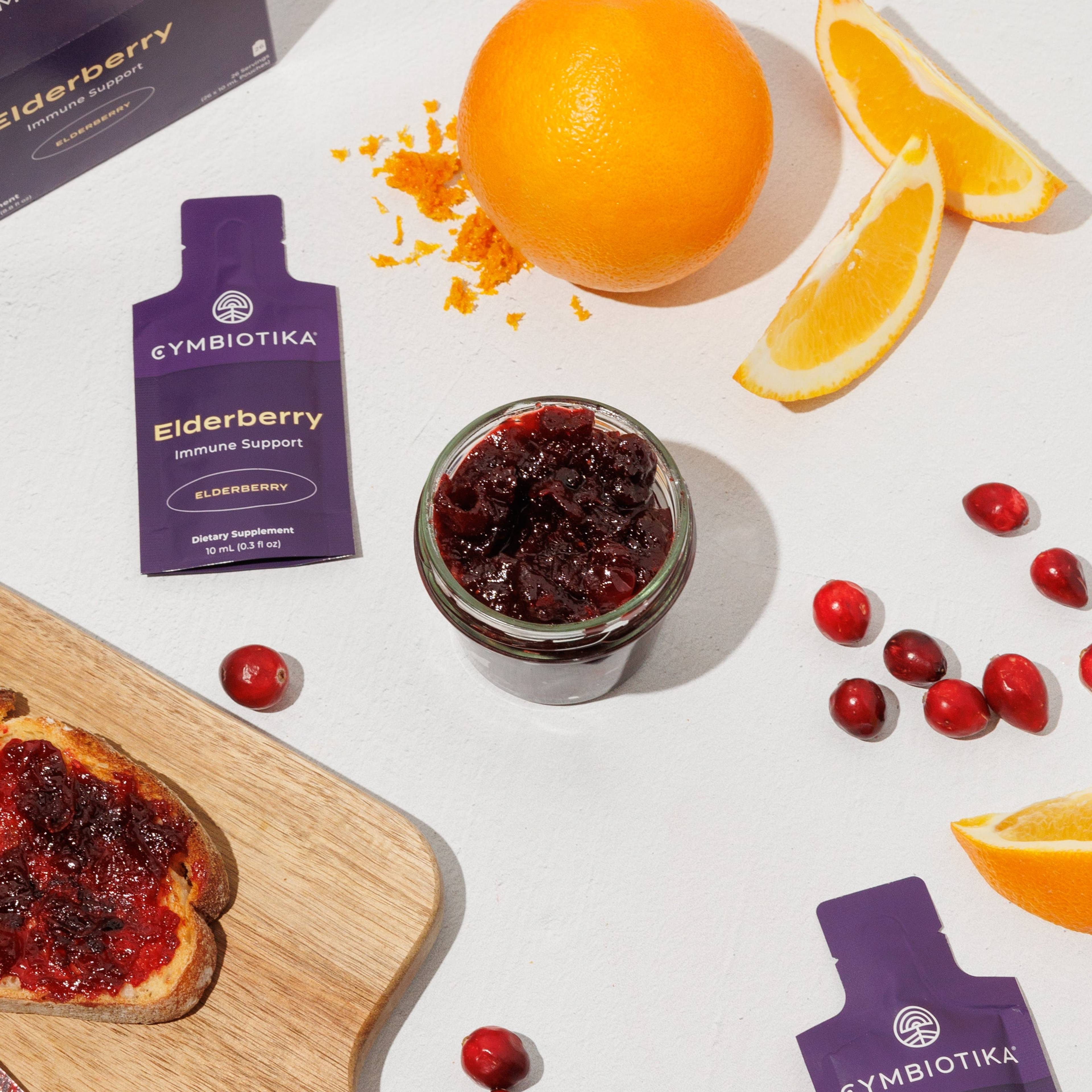 Elderberry supplement packets beside cranberry jam, oranges, cranberries, and toast on a cutting board.
