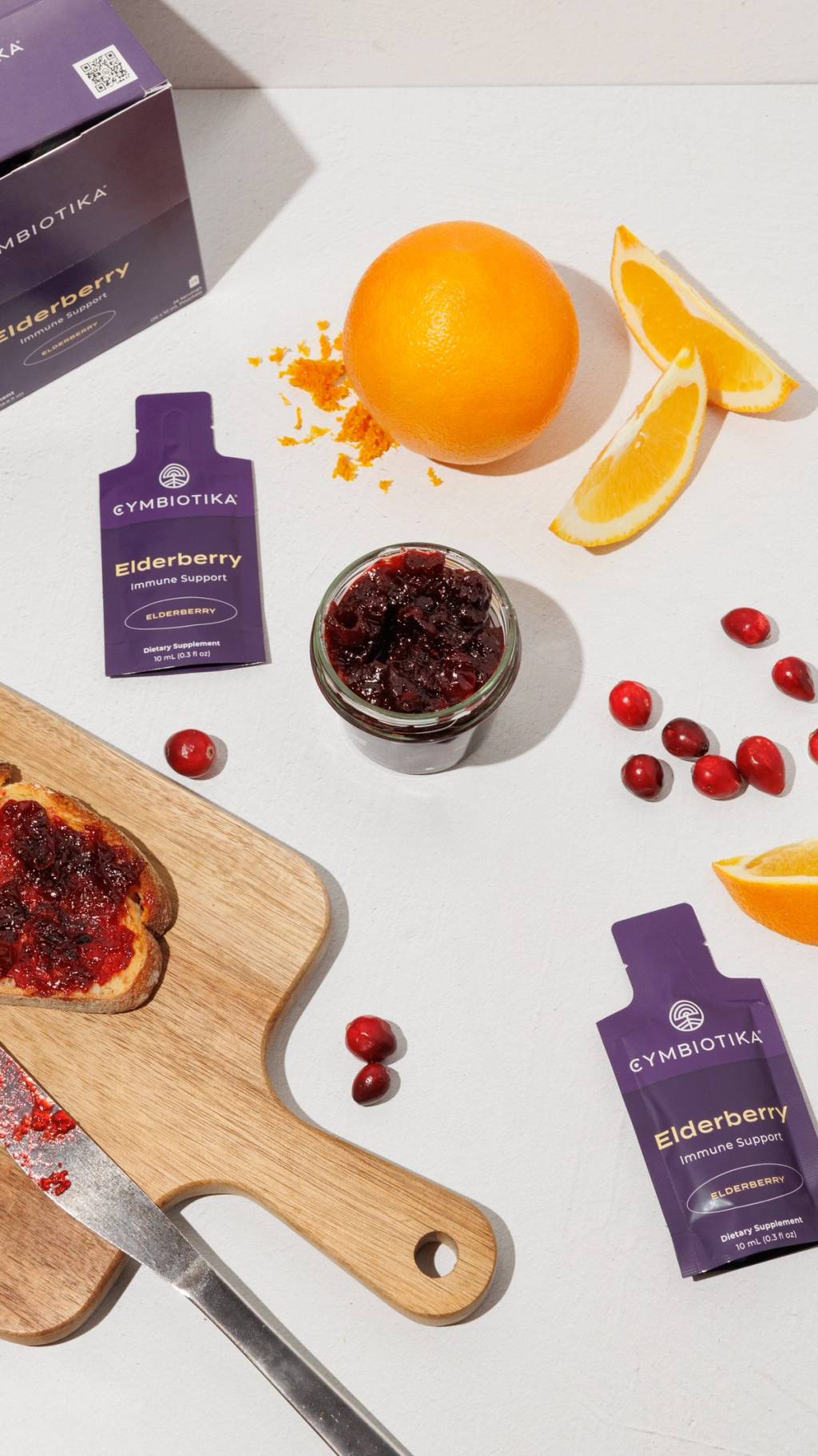 Elderberry supplement packets beside cranberry jam, oranges, cranberries, and toast on a cutting board.