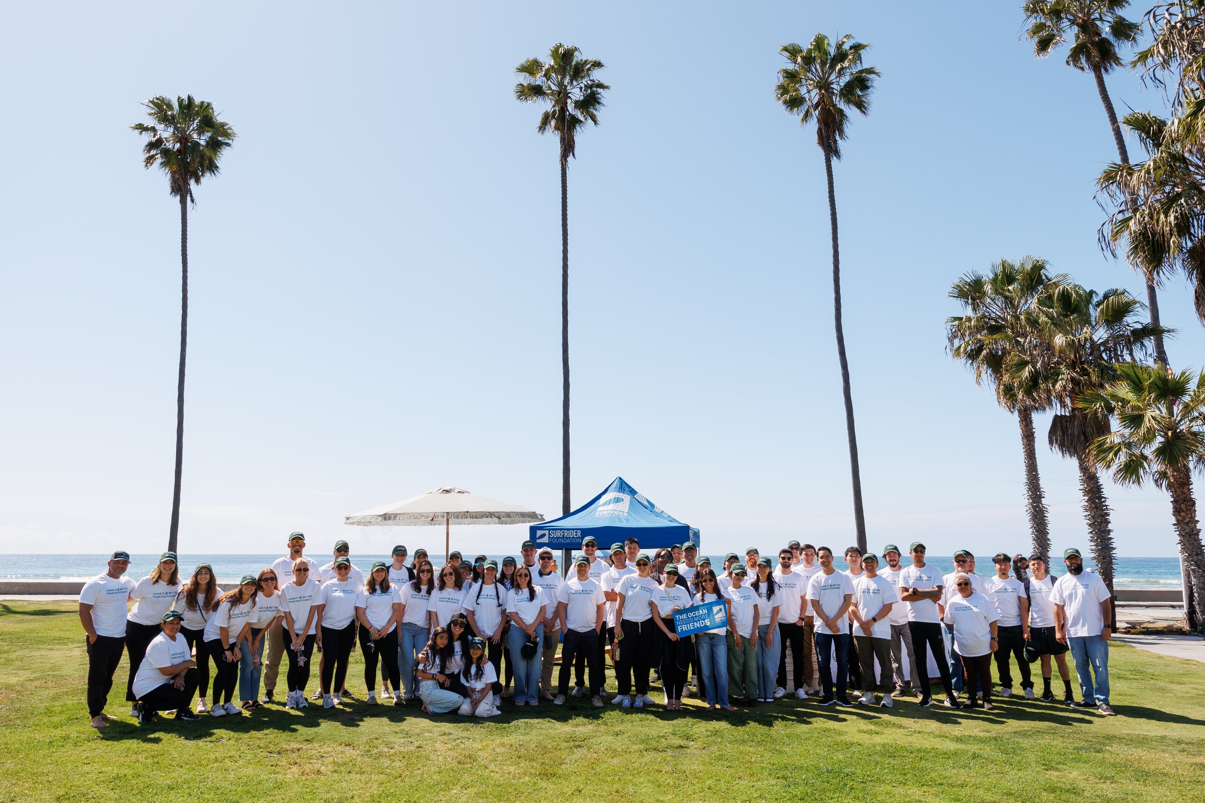 Large group in white shirts posing on a grassy beachfront beneath tall palm trees.