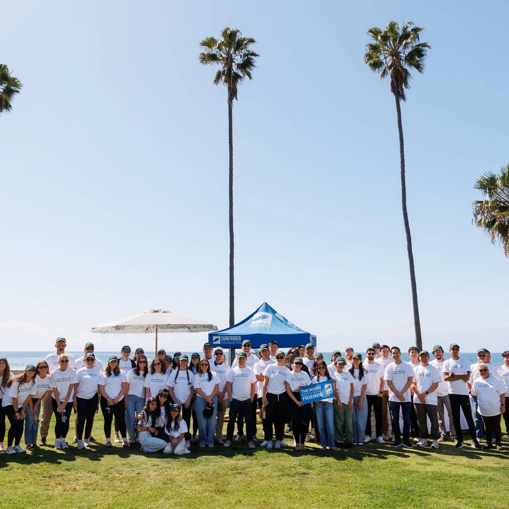Large group in white shirts posing on a grassy beachfront beneath tall palm trees.