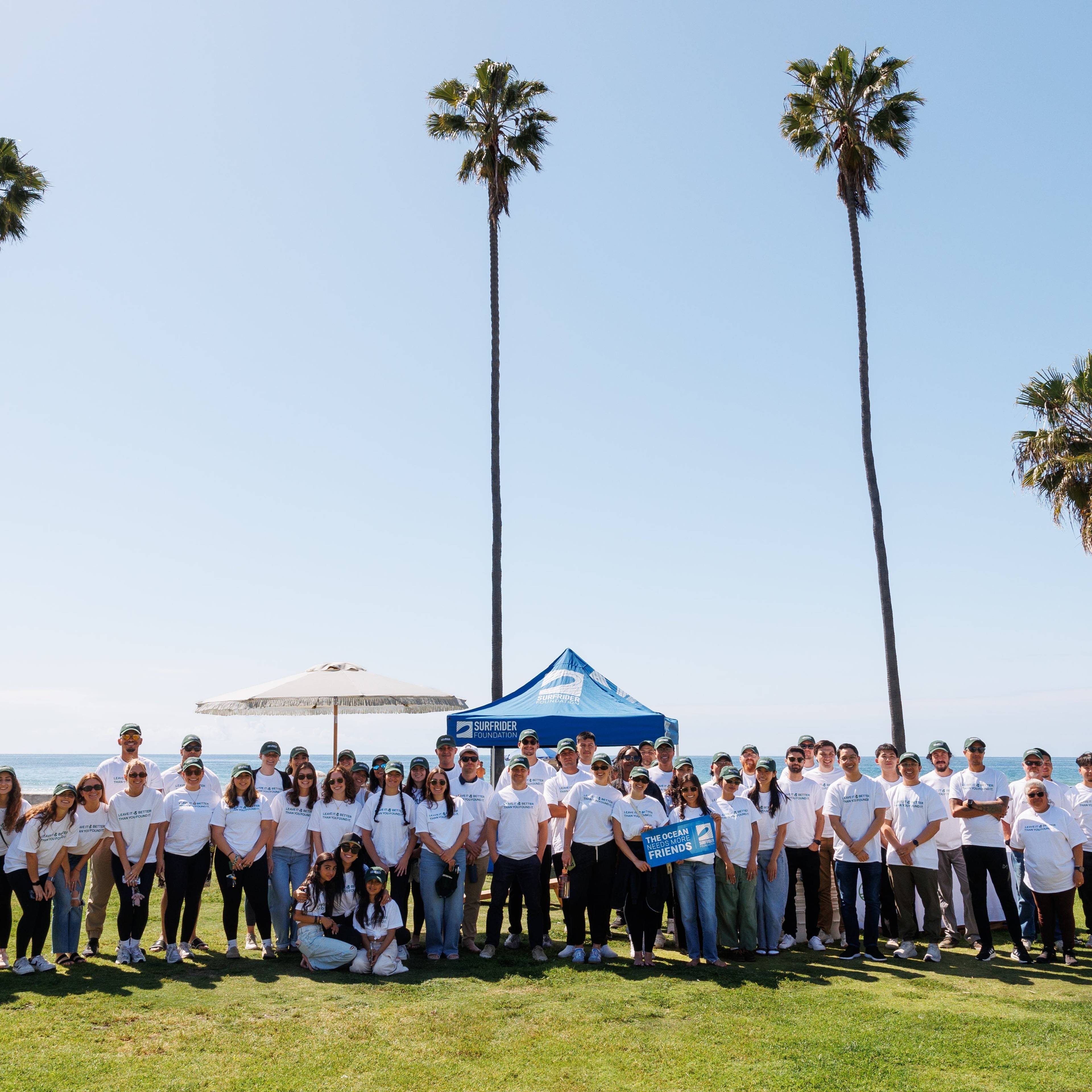 Large group in white shirts posing on a grassy beachfront beneath tall palm trees.