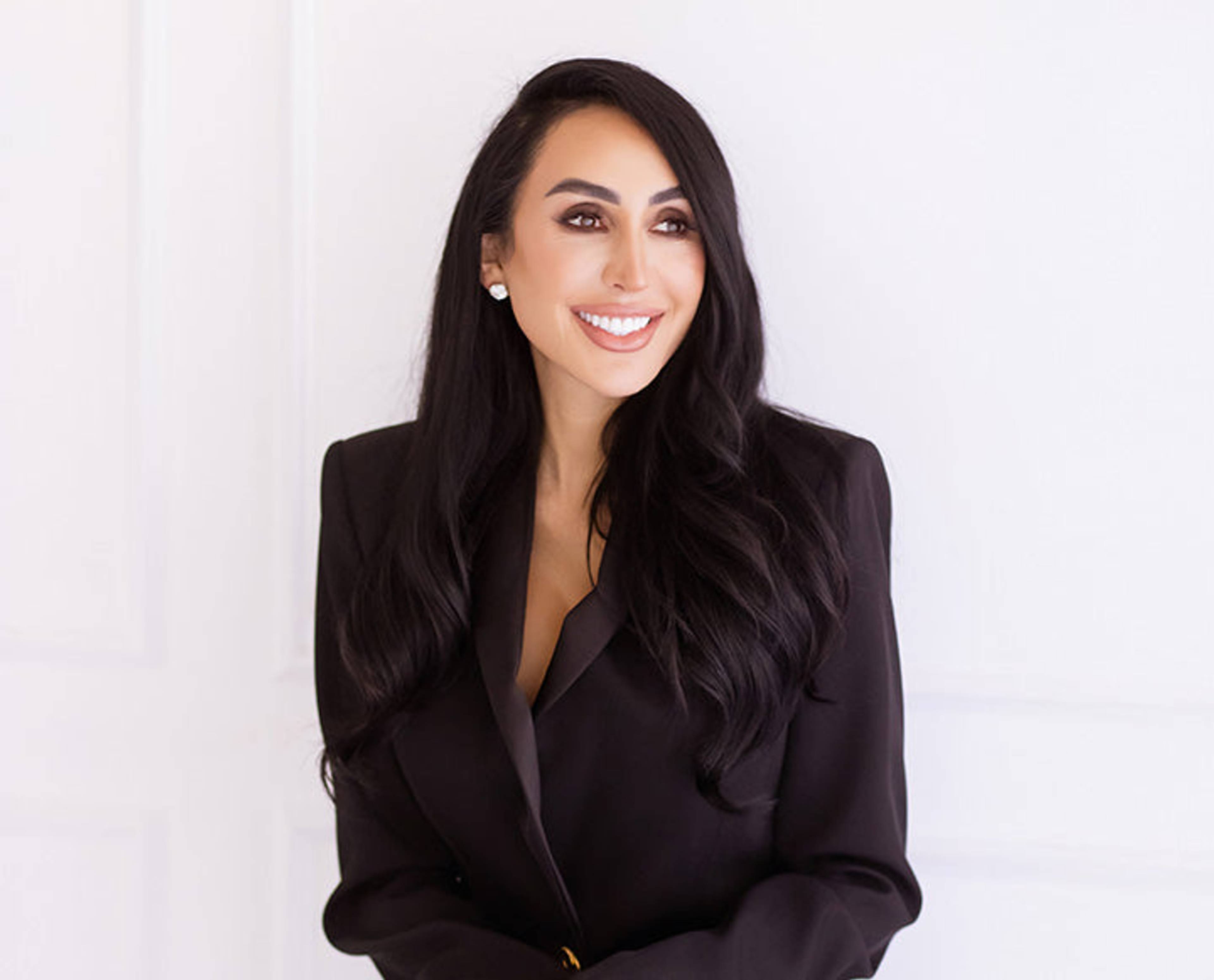 Smiling woman with long dark hair wearing a black blazer against a white wall.