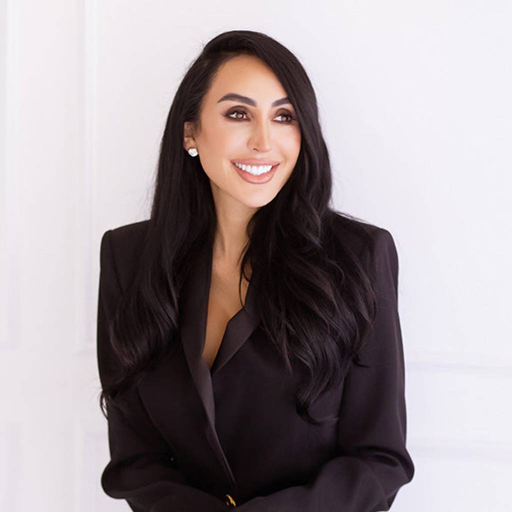 Smiling woman with long dark hair wearing a black blazer against a white wall.