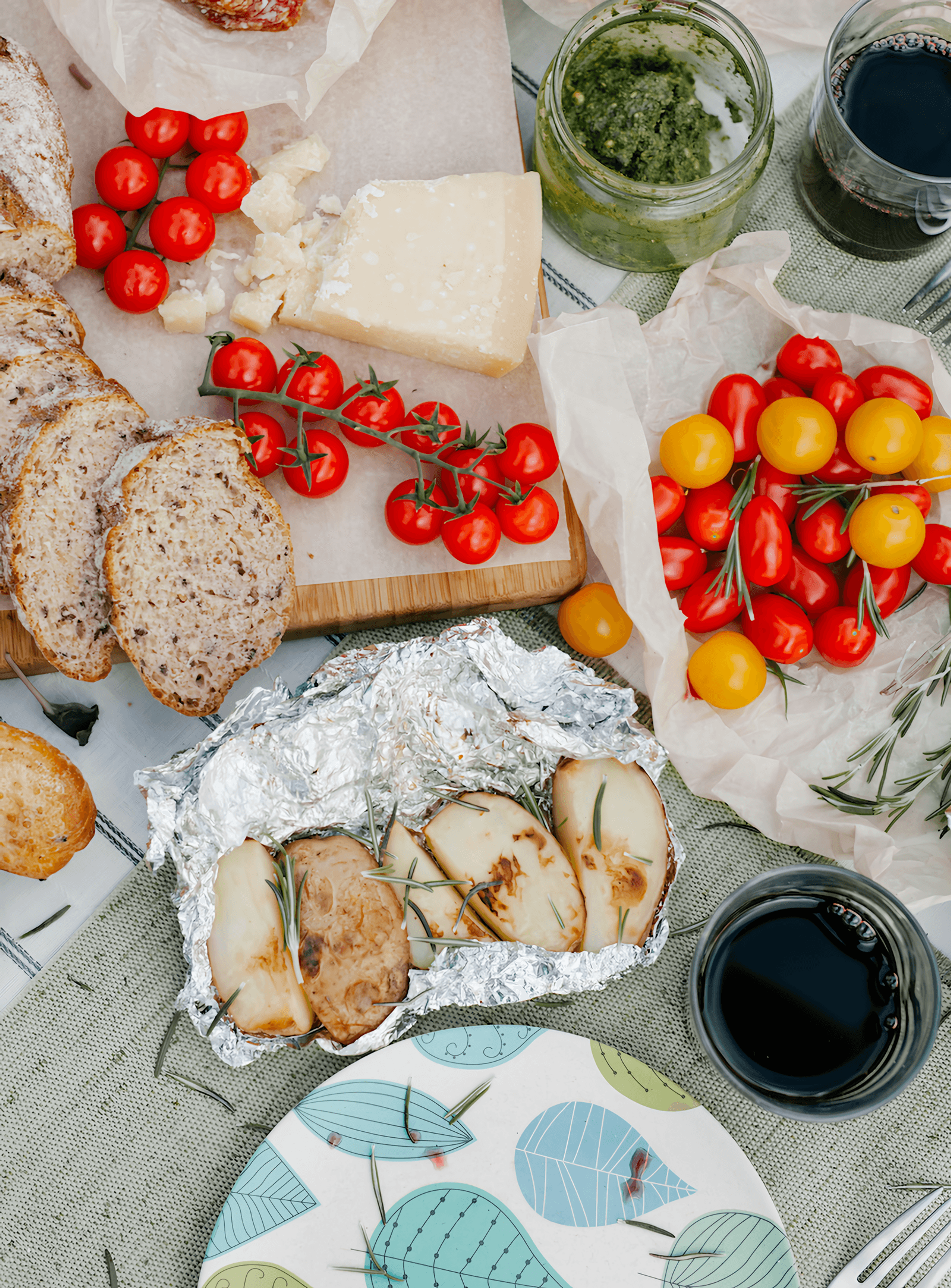 Overhead view of bread, tomatoes, cheese, pesto, and roasted food wrapped in foil, with dark drinks on a table.