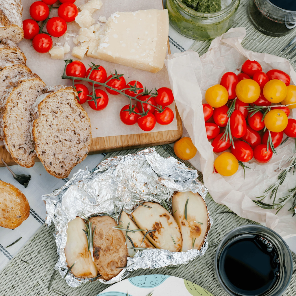 Overhead view of bread, tomatoes, cheese, pesto, and roasted food wrapped in foil, with dark drinks on a table.