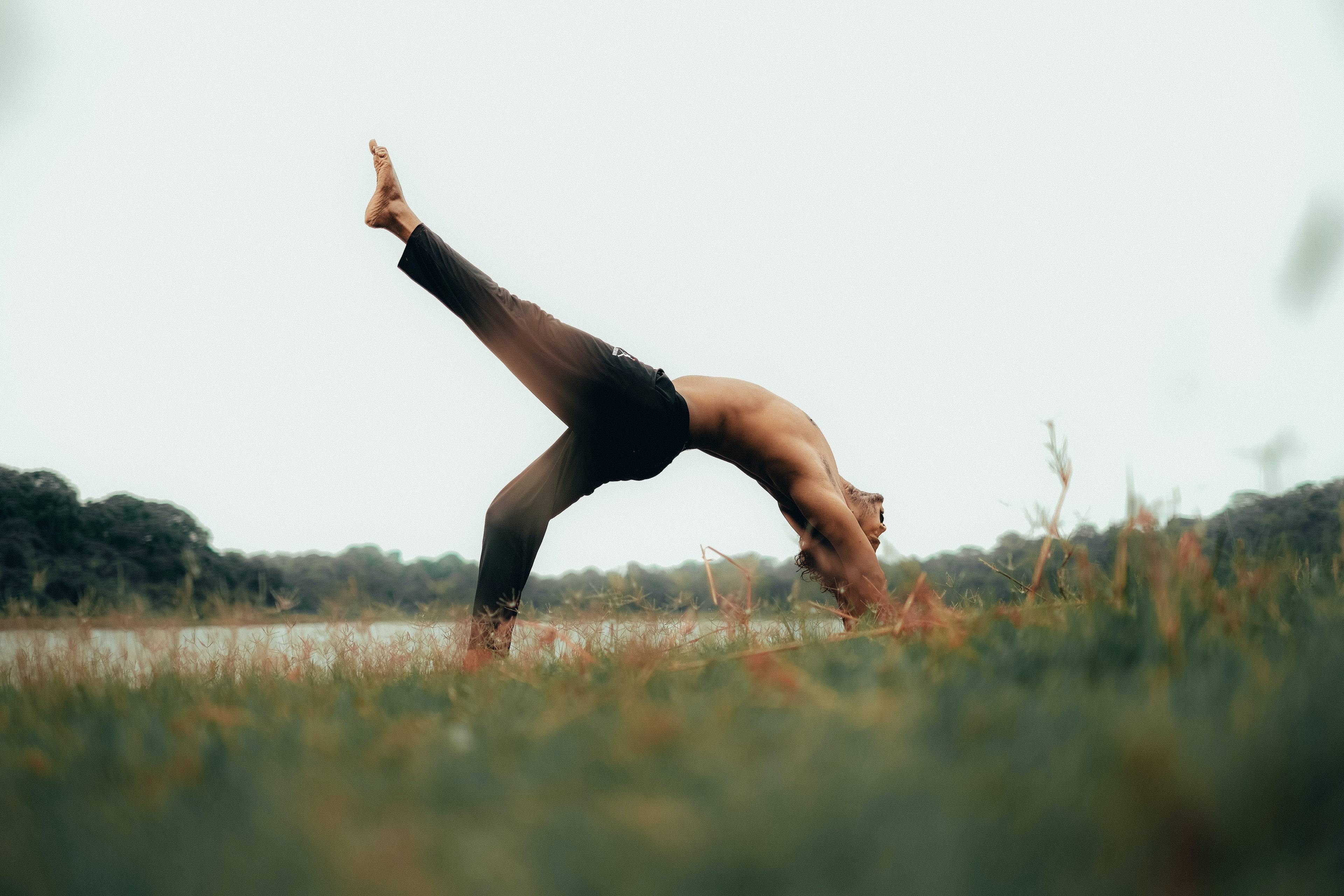 Shirtless man practicing a one-legged backbend outdoors beside a lake.